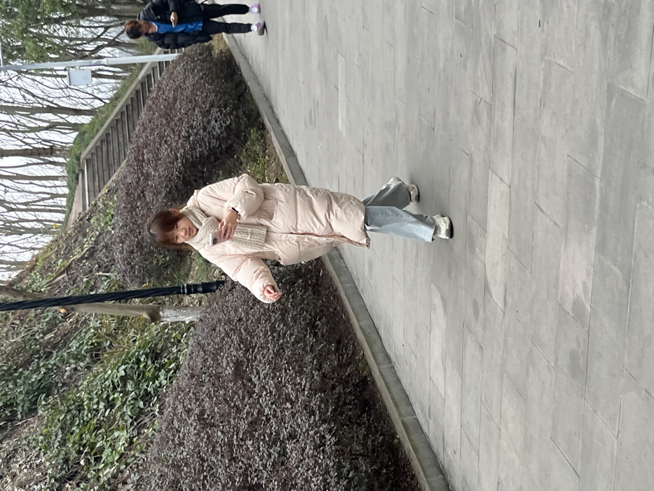 A woman in a long, light-pink puffer coat, blue jeans, and white sneakers walks on a gray paved path. She is holding a phone and wearing a light-colored scarf. Behind her, another person in a black jacket and dark pants is also walking on the path. The path is bordered by bushes on one side and a slope with trees and stairs on the other. A pole is seen on the left side of the image.