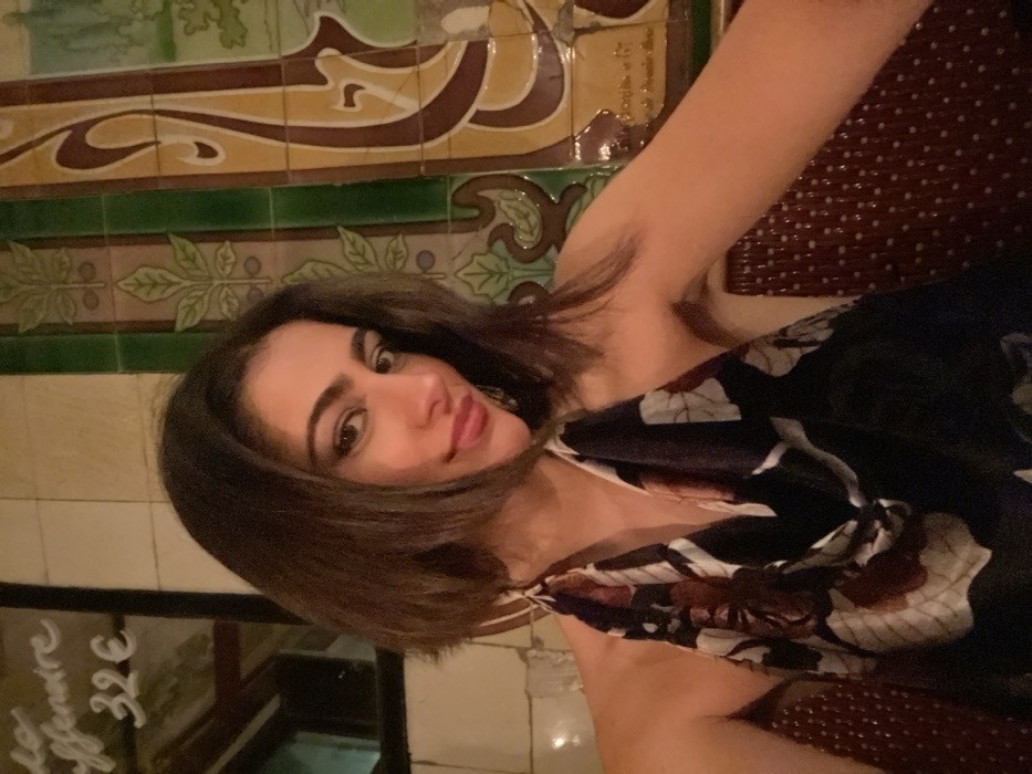 A woman with shoulder-length brown hair and fair skin poses indoors. She wears a floral print dress, exposing one shoulder. Behind her, decorative tiles with brown and green patterns line the wall. To her left, a chalkboard displays writing in a light color, including the price "32€". A woven, dark red surface is visible in the upper right.