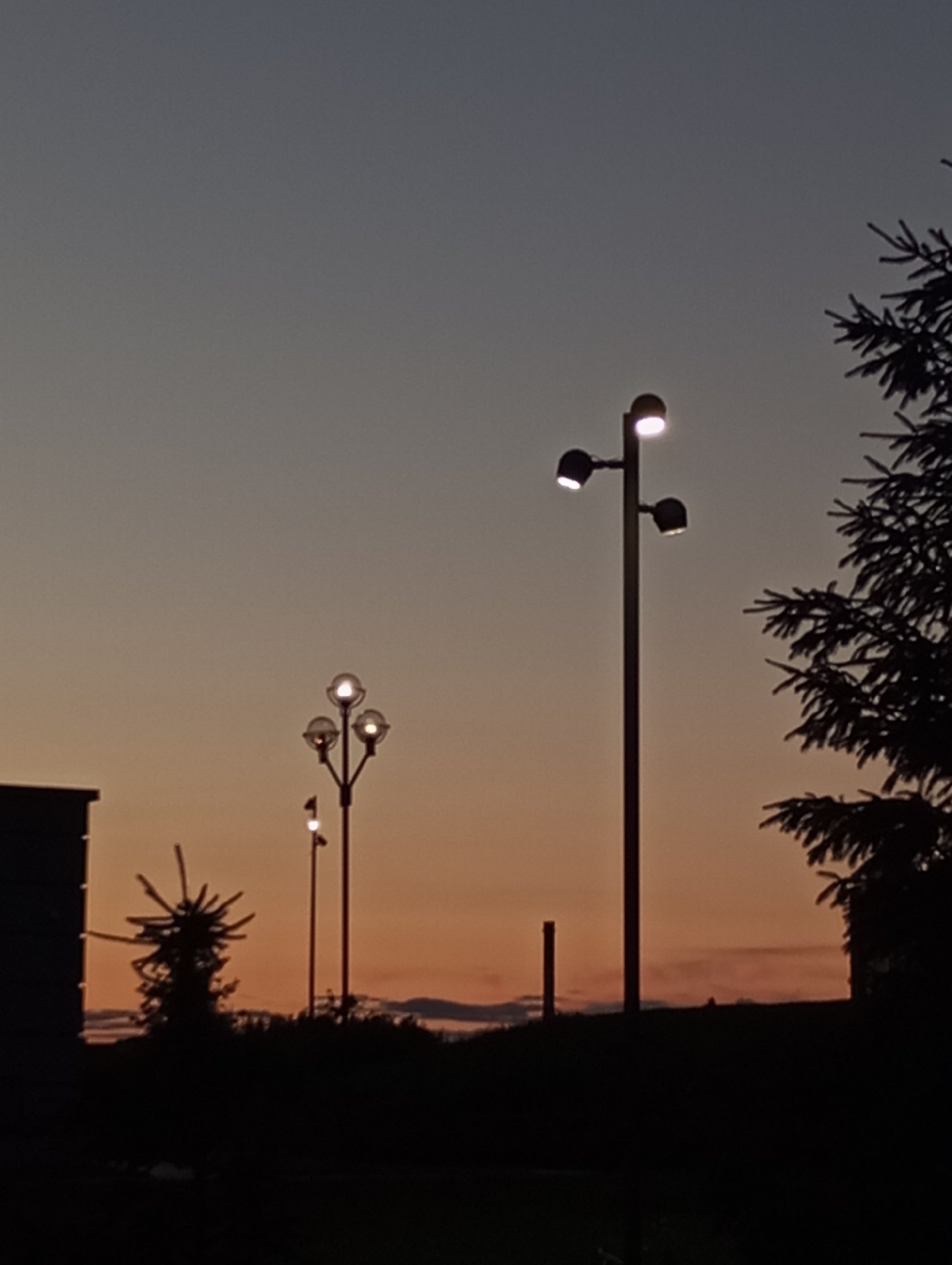 The image captures a dusk or dawn scene, likely in Pervomaysky, Russia, given the prompt. The sky exhibits a gradient of colors, transitioning from a dark grey/blue at the top to a warmer orange/peach near the horizon. Silhouetted elements dominate the foreground. Several lampposts of varying designs are visible. One, near the right, features a tall pole with three directional lights. Another has a more classic design with globe-shaped lights, likely a cluster of three. A much smaller single lamppost is placed between the building on the left and the globe-shaped lamps. A building is partially visible on the left edge of the frame, appearing as a dark silhouette. Silhouetted trees and bushes form the landscape, with one tree visible on the right side. A small chimney is seen to the left of the most right lamppost.