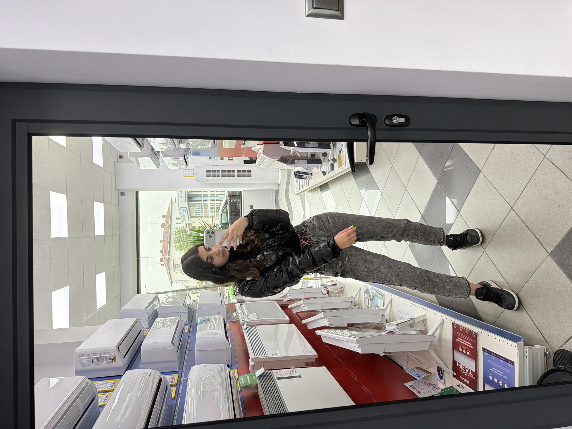 A long shot shows a reflection of a young woman taking a photograph of herself inside a retail store located in Unknown, Unknown. The woman is standing behind a low counter displaying rows of HVAC units and wearing a black, puffy jacket and gray pants with black shoes. The woman has dark hair. The floor is tiled in a light gray, diamond pattern. In the background, a storefront can be seen.