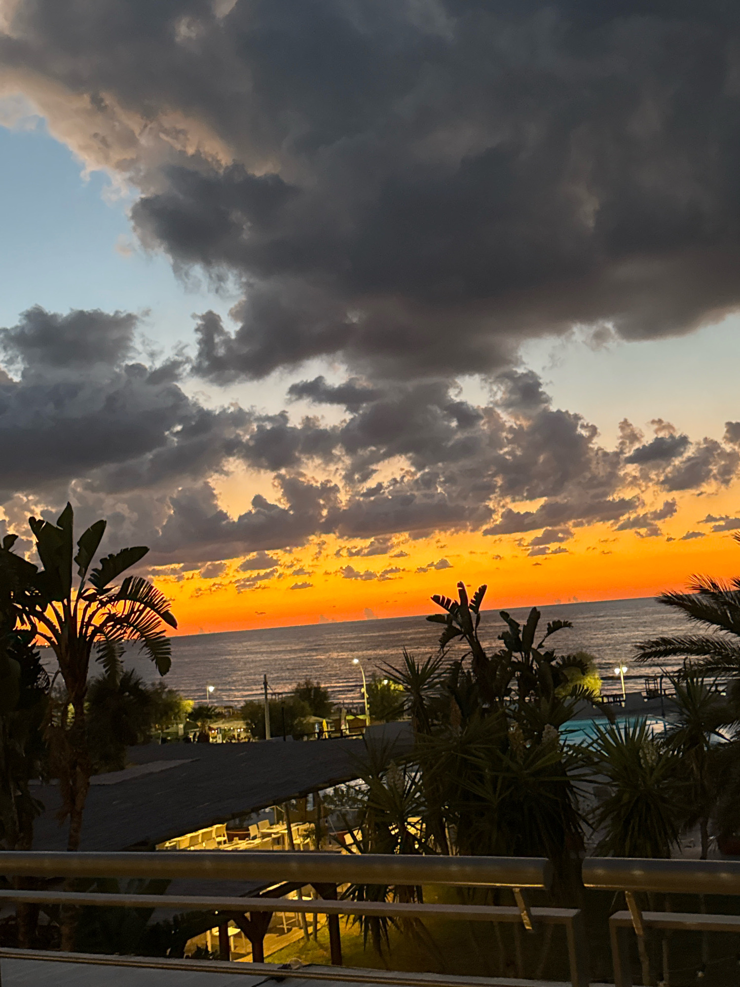 The image depicts a sunset over the sea in Isola delle Femmine, Italy. Dark clouds dominate the upper portion of the sky, contrasting with a vibrant band of orange and yellow light near the horizon, indicating the setting sun. The sea is visible below the colorful sky, and the coastline is lined with palm trees and other vegetation. In the foreground, there appears to be a building with tables and chairs, possibly a restaurant or outdoor seating area, and a railing from a balcony or walkway. The scene is likely captured during the evening.