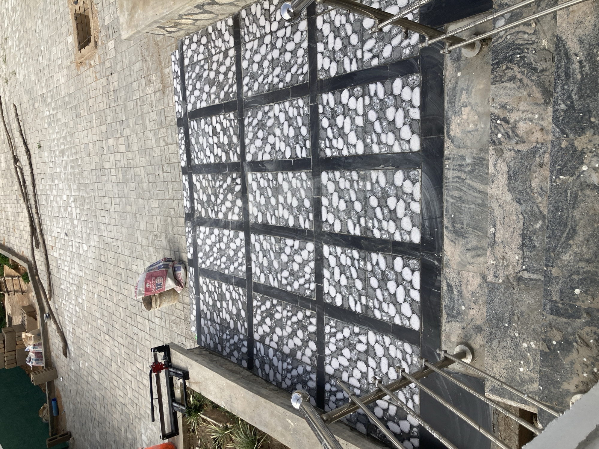 The image shows a view from above, possibly a balcony or elevated position, of an outdoor area in Jos, Nigeria. The ground is covered with various types of paving. One section features a black and white patterned tile arrangement, made up of 12 square sections with black lines separating them. Each square is filled with an abstract design of rounded shapes. Adjacent to this is a set of stairs with a marble-like pattern and a metal railing.
To the left of the tiled area is a brick-paved surface. On this surface, a bag, possibly containing cement or other building materials, is visible, along with what appears to be a tile cutter. Some bricks, likely building supplies, are near a patch of artificial turf. Long wooden poles and other construction materials are also present.
