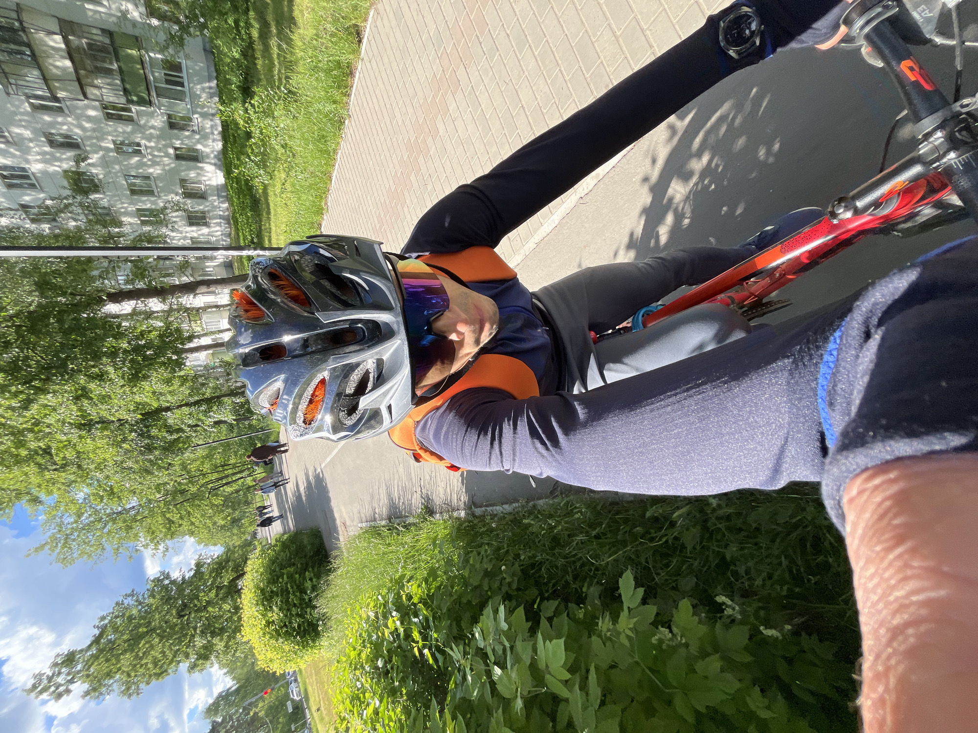 A cyclist wearing a helmet and sunglasses is riding a bicycle on a paved path. To the left, a grassy area with trees lines the path, beyond which is a multi-story building. On the right is more greenery. The cyclist's arm and wristwatch are visible, holding the bicycle's handlebars. People are walking in the distance on the path. The photograph was taken on a sunny day in Chaikovsky, Russia.