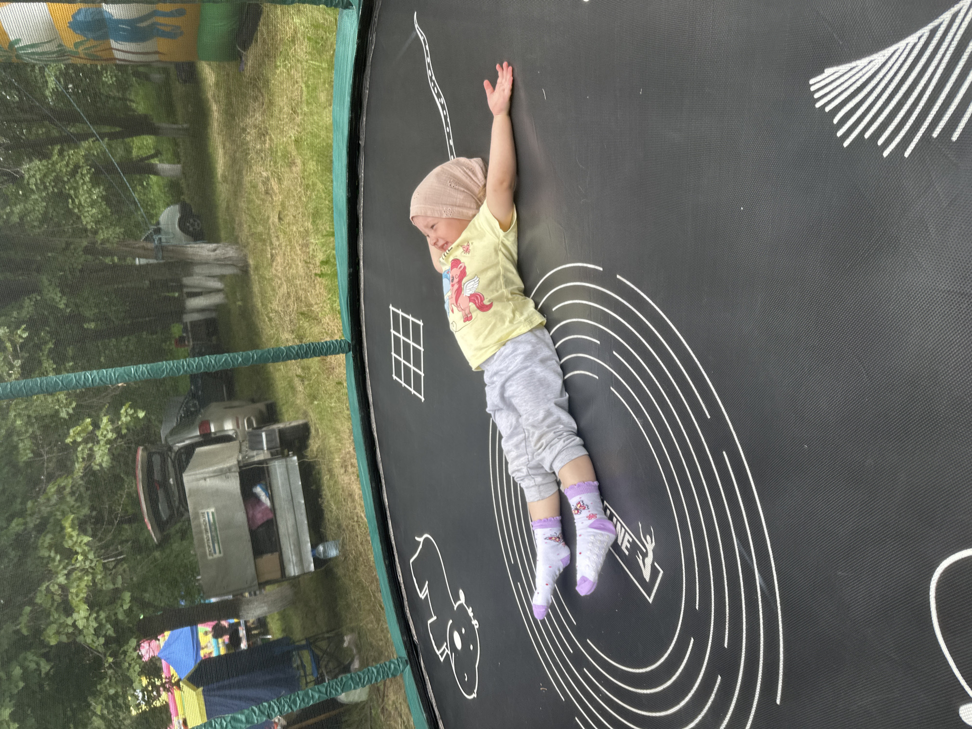 A young child lies on their back on a black trampoline with white markings. The child wears a light brown head covering, a yellow t-shirt with a pink winged creature graphic, grey sweatpants, and white socks with purple trim and small pink flowers. The child's right arm is extended to the side, palm up, and their left arm is bent at the elbow, resting near their head. The trampoline is in an outdoor setting with trees and grass visible in the background. To the left of the trampoline, there's a blue and yellow inflatable structure and a silver trailer attached to a vehicle. Further in the background, other vehicles and trees are present. The scene appears to be within a park or recreational area, possibly at a festival or event. The location is specified as Yaroslavka, Russia.