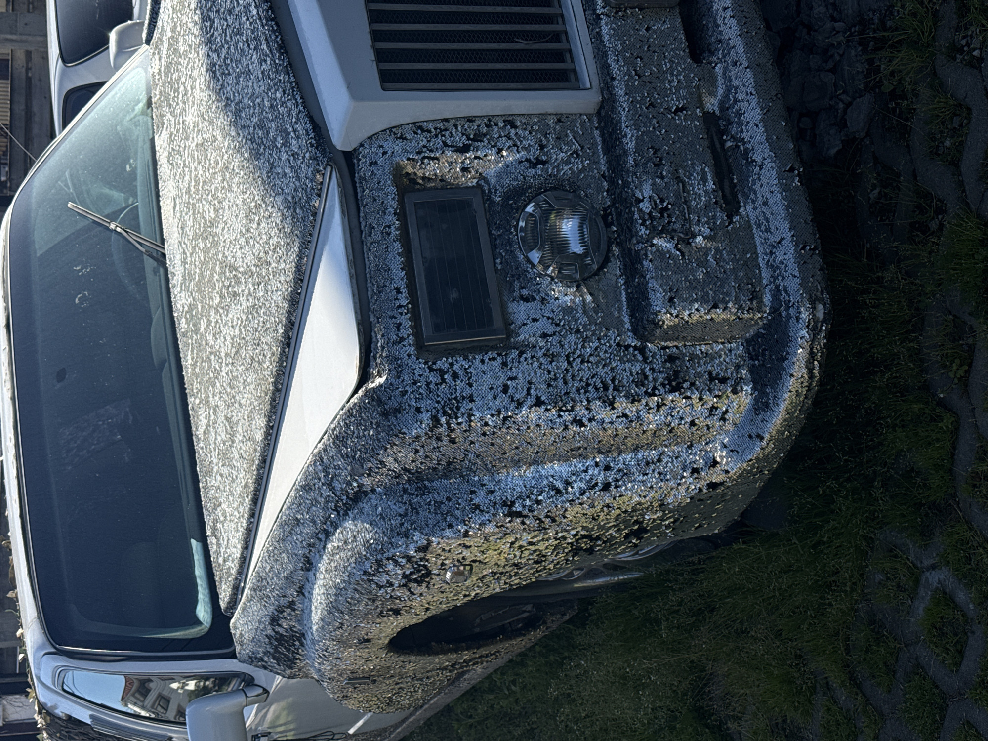 The image displays the front and side view of a car parked outdoors in Sochi, Russia, during bright daylight. The vehicle's most distinctive feature is its exterior, which is almost entirely covered in small, shiny silver sequins, giving it a highly reflective and shimmering appearance.
The car's hood, front bumper, and a portion of its side are visible, all adorned with these sparkling sequins. On the front, a black rectangular panel (possibly a vent or small solar panel) and a round, textured light fixture are integrated into the sequined surface. Part of the windshield and a single wiper blade are also visible. The car's underlying color appears to be light, possibly white or light grey, beneath the sequin embellishment.
To the right and foreground of the car, there is a patch of green grass interspersed with grey concrete paving blocks, indicating a parking area or roadside setting. The strong sunlight creates significant glinting highlights on the sequins, suggesting clear weather conditions. No people are visible, and there is no discernible text in the image.