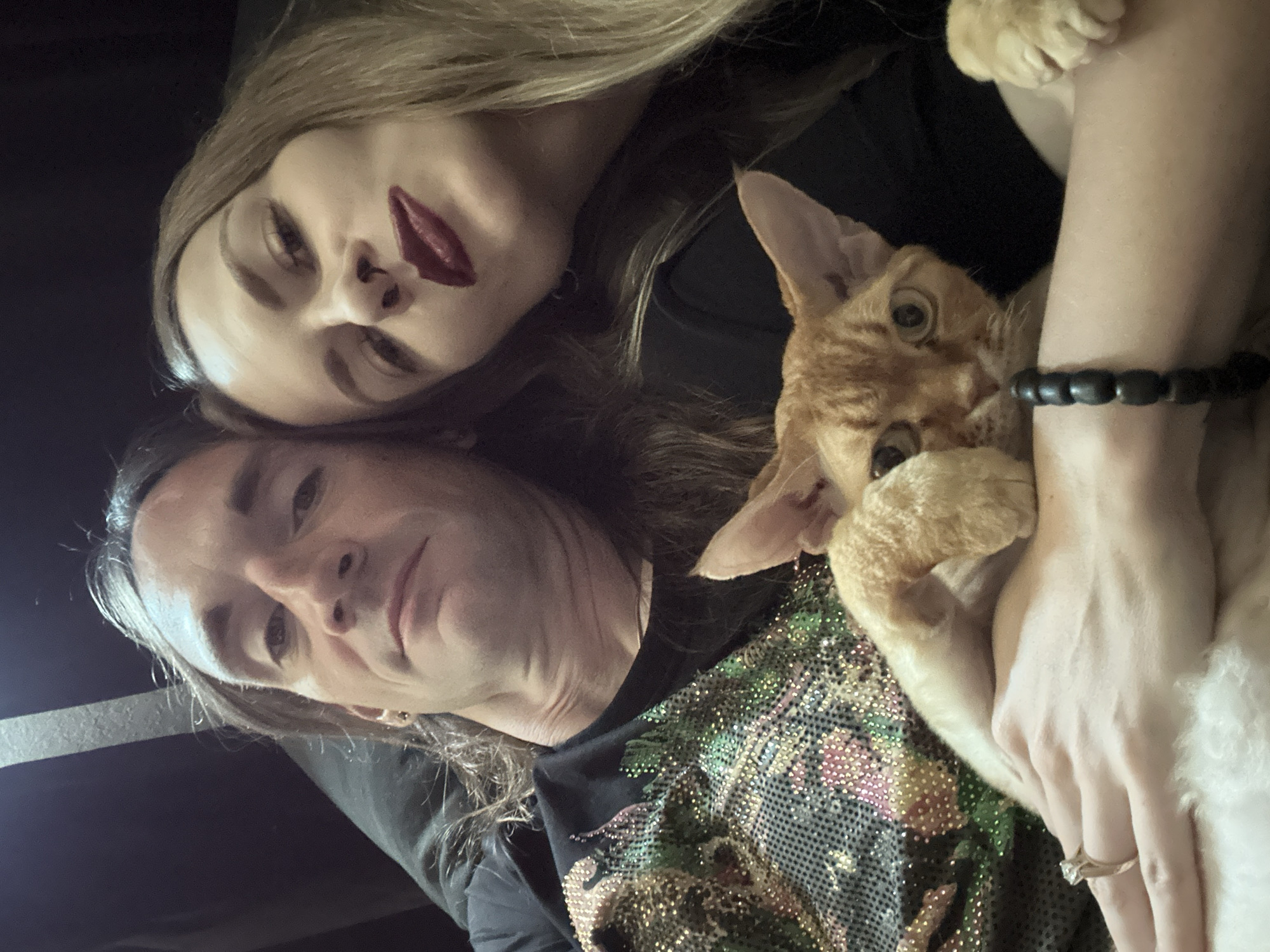 The image shows a selfie taken by a man, positioned in the lower left quadrant of the frame. A woman, identified by her long blond hair and distinctive dark red lipstick, is above him. An orange cat is nestled in the arms of the woman. The man is wearing a black shirt with an ornate, possibly floral, pattern rendered in what appears to be sequins or rhinestones. A bracelet is on the woman's wrist. The environment is dark and featureless, offering no clues about the location within Cape Coral, United States of America. The action in the image is a group selfie.
