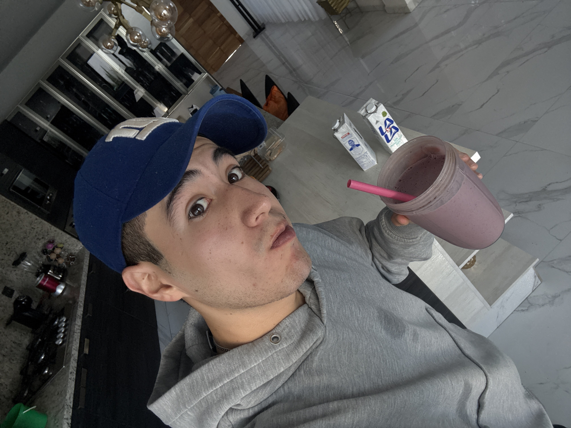 A young man wearing a blue baseball cap and a gray hoodie holds up a purple smoothie with a pink straw. Two cartons of milk stand on a light-colored counter behind him. He appears to be indoors, possibly in a kitchen or dining area. The location is likely San Bernardino Tlaxcalancingo, Mexico, although this is not directly discernible from the image itself.