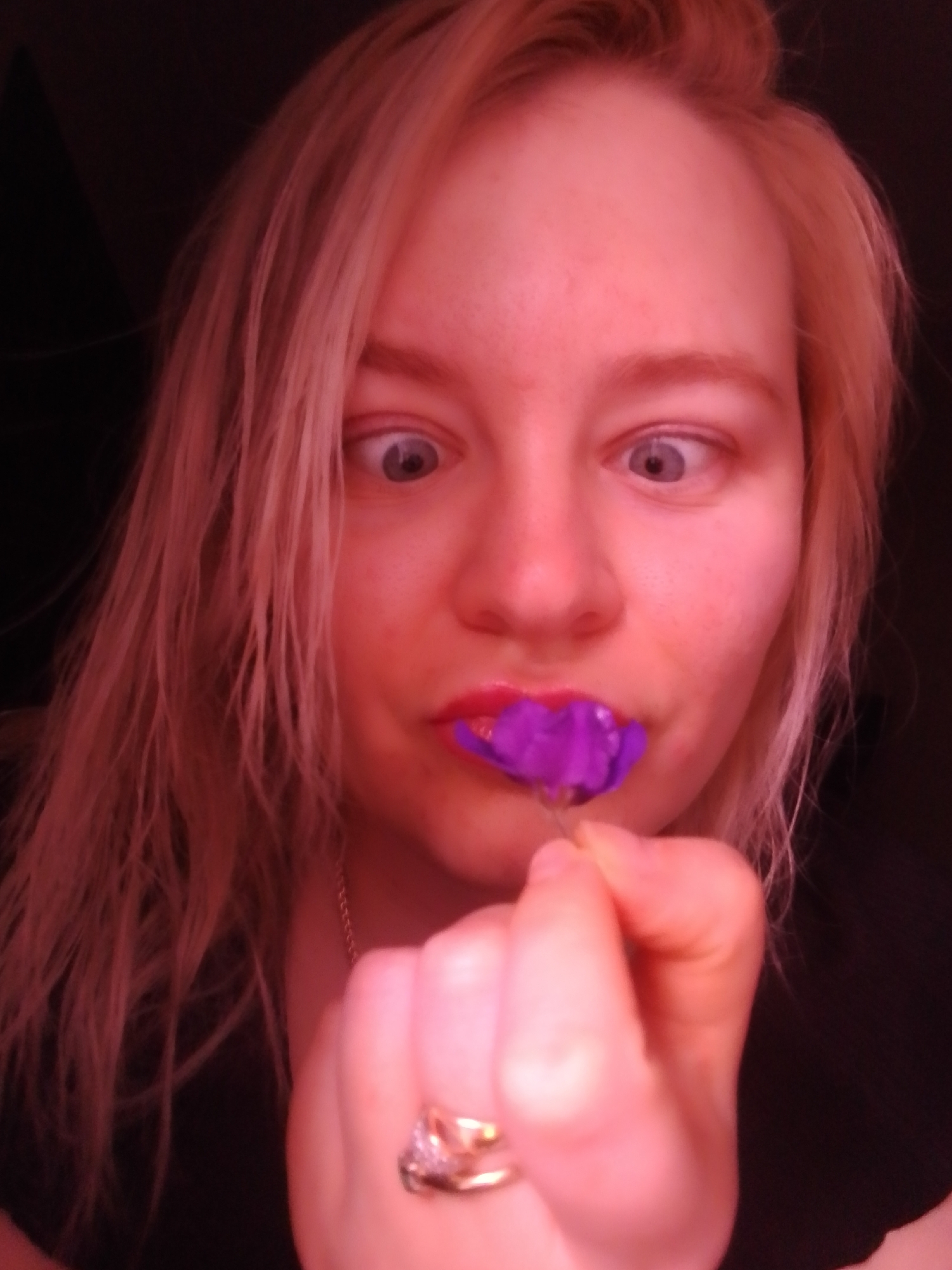 A woman with light-colored hair holds a purple flower to her lips while looking wide-eyed at the camera. She is wearing a ring on her finger and a necklace. The background appears dark. It is not possible to determine her location as Izhevsk, Russia based on visual information.