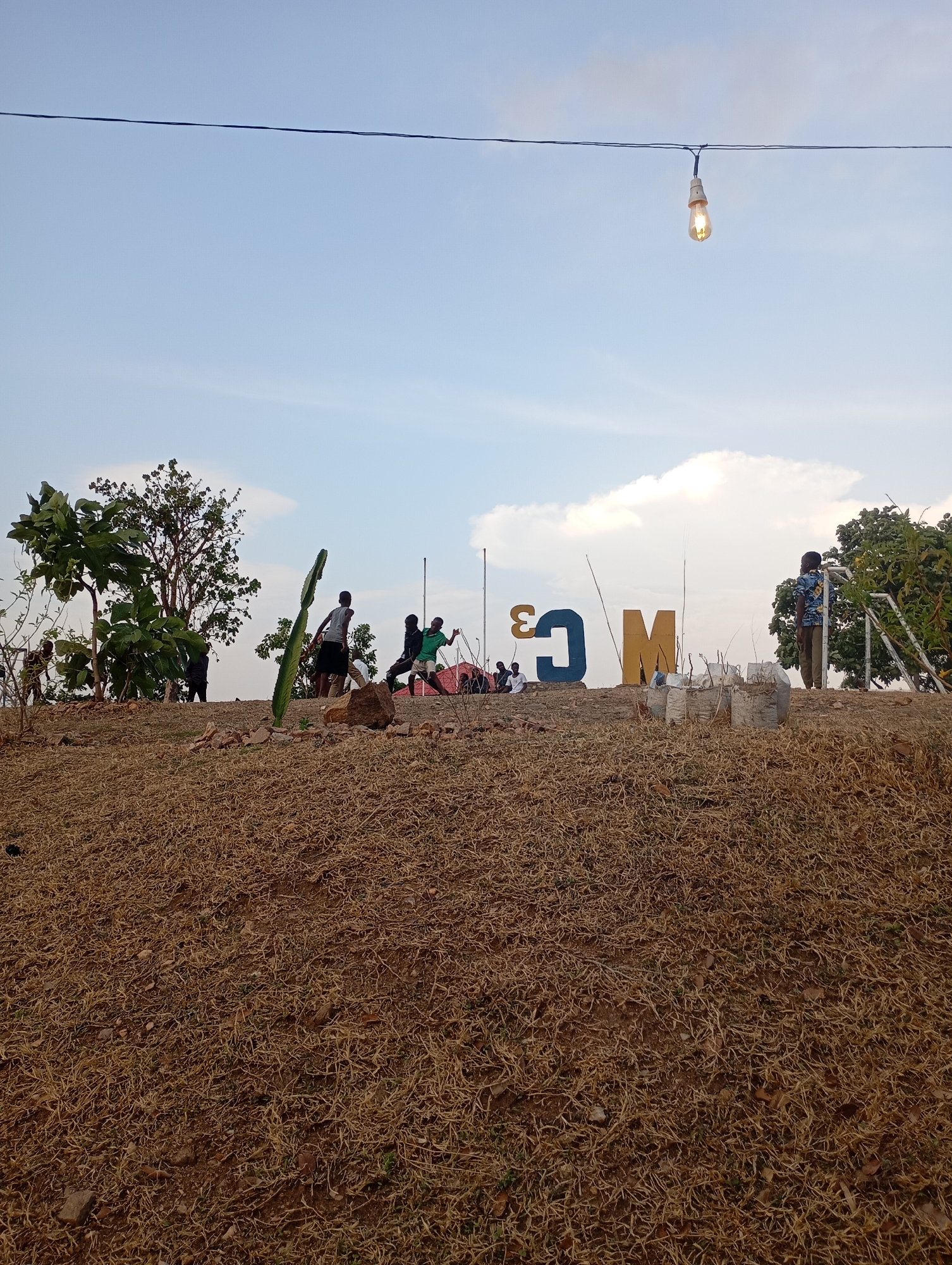 The image shows a grassy hilltop in Uke, Nigeria. Several people are on the hill; some appear to be moving around, while one stands near the edge. Large, blue and yellow letters spelling "COM" are erected on the hill. Plant life, including trees and possibly a cactus, are visible. An unlit lightbulb hangs suspended from a wire strung across the sky. The ground is covered in dry grass.