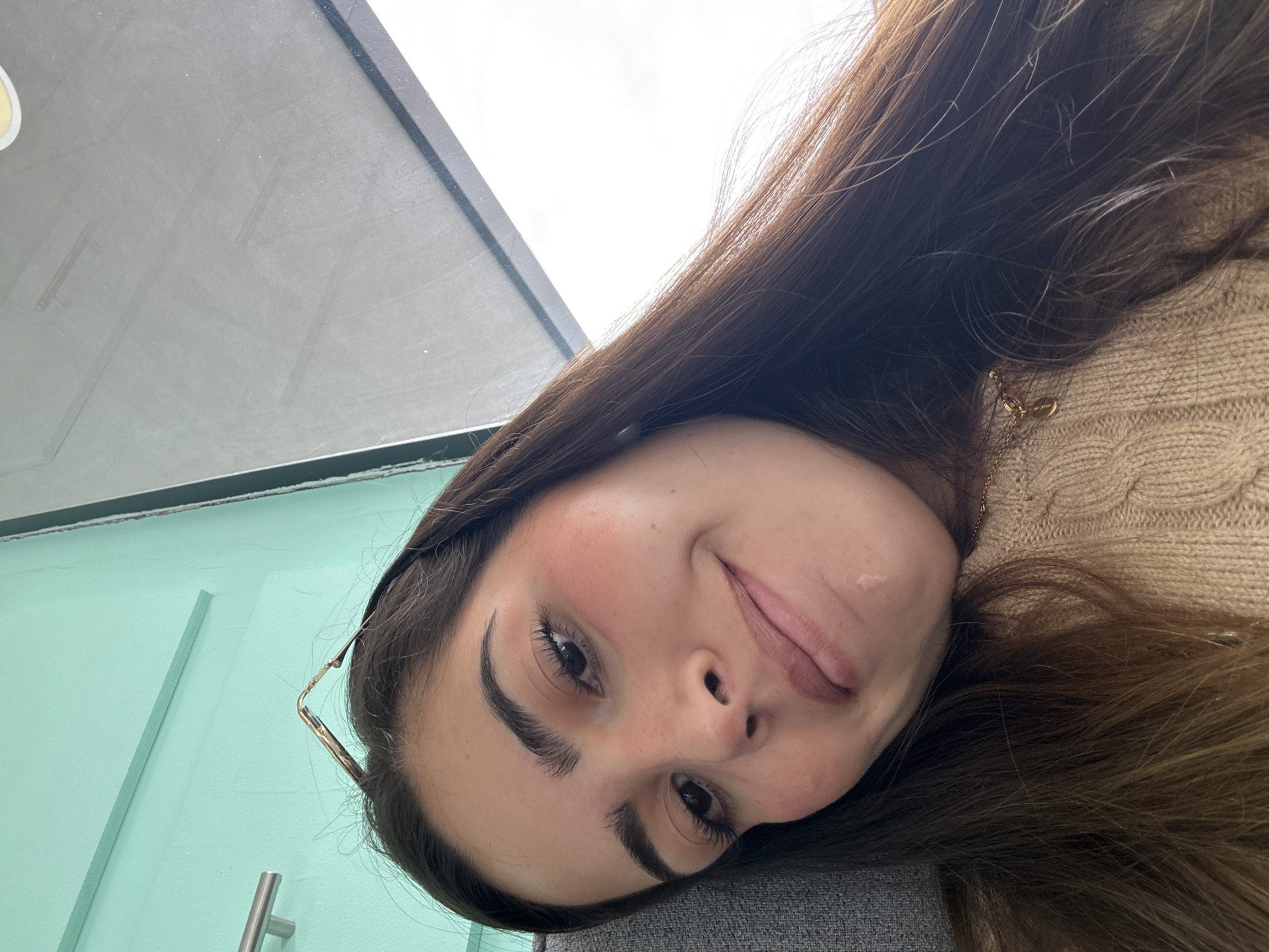 A young woman is featured in a close-up, rotated selfie, displaying a playful and casual expression with her tongue slightly visible. Her long, dark brown hair flows around her face. She has prominent eyebrows, dark eyes, and a subtle blush on her cheeks. She is wearing a light beige or tan knitted sweater, and a delicate gold chain necklace with a small, indistinct pendant is visible. A portion of gold-rimmed eyeglasses is seen resting in her hair on the left side of the frame.
The setting appears to be indoors during the day, with an overcast sky visible through a window or skylight in the upper left. A dark grey or black frame separates this opening from a light blue-green wall below, which features a vertical molding and a silver-colored handle. The woman's head rests on a dark grey, possibly upholstered surface. The overall mood is relaxed and lighthearted. No discernible text is visible in the image. This scene is located in San Bernardino Tlaxcalancingo, Mexico.