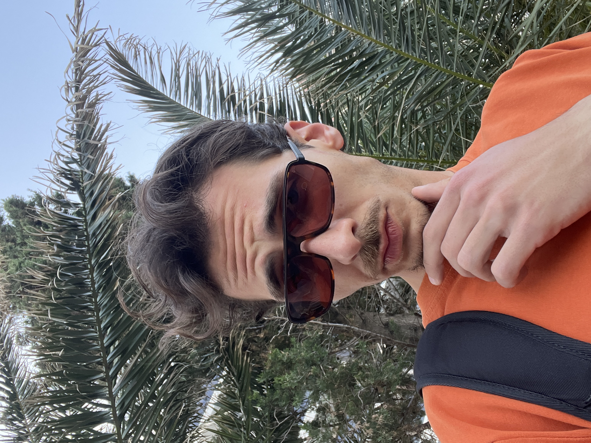 The image, originally captured in portrait orientation but presented rotated 90 degrees counter-clockwise, features a close-up outdoor portrait of a young adult man. He is looking directly at the camera with a focused or contemplative expression, with visible wrinkles on his forehead. He wears dark sunglasses with a tortoiseshell-patterned frame and an orange short-sleeved top. A black strap, likely from a bag or backpack, is visible over his right shoulder (when oriented upright). His left hand is resting gently against his chin and neck area. He has dark, wavy hair and a short beard/stubble with a mustache.
The background is a vibrant outdoor scene under a clear blue sky. Dense green foliage, primarily large palm fronds (some appearing dry or lighter in color) and other evergreen trees and bushes, fills the rest of the frame. The bright natural light indicates it is daytime with clear weather. There is no visible text in the image. The presence of palm trees and lush greenery is consistent with the provided location context of Budva, Montenegro.