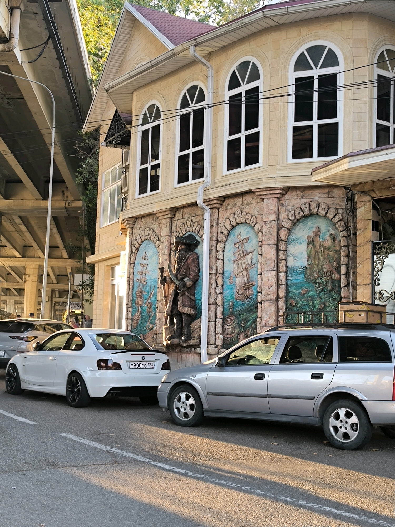 The image shows a street scene in Sochi, Russia. In the foreground, two cars are parked on the side of the road. The car on the left is a white BMW coupe, and the car on the right is a silver station wagon. Behind the cars, there is a building with a yellow facade, arched windows, and decorative murals on the lower level. One mural depicts a pirate figure, and others show sailing ships. Above the building, a concrete overpass can be seen, with trees and foliage peeking through. The scene is illuminated by the warm light of what appears to be late afternoon or early morning.