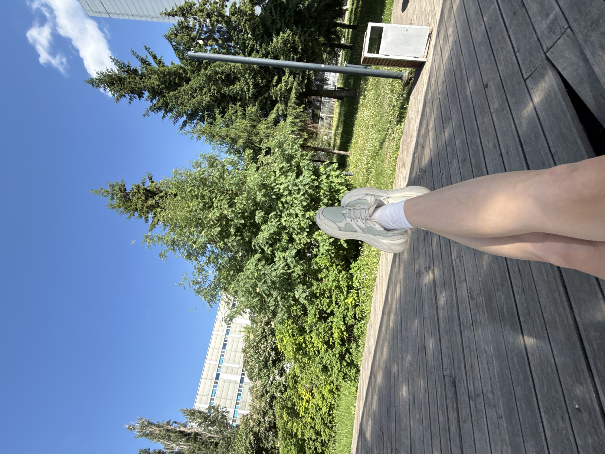 The image shows a person seated on a wooden boardwalk, likely outdoors, with their legs crossed. The person is wearing white socks and light-colored sneakers. The boardwalk is made of wooden planks, and sunlight creates a pattern of light and shadow on its surface. The background features lush greenery with various trees and foliage. A building is visible in the distance. The sky is bright blue with scattered clouds. A trash receptacle is on the boardwalk to the left of the legs and greenery. Given the presence of trees and the modern architecture, this scene could be found in Kazan, Russia.