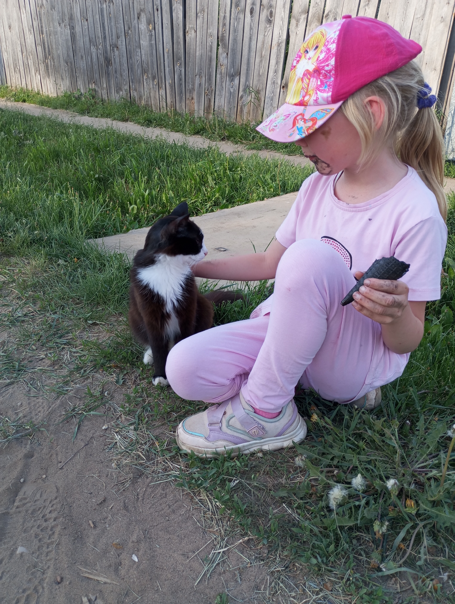 A young girl wearing a pink shirt, pink pants, and lavender sneakers, is sitting on the ground next to a black and white cat. She is wearing a pink baseball cap with a cartoon character on it. The girl has dark smudges around her mouth and appears to be eating a dark-colored ice cream cone held in her right hand. She is petting the cat with her left hand. The scene appears to be outdoors, near a grassy area and a dirt patch, with a wooden fence in the background. The location is in the city of Буренка, Russia.