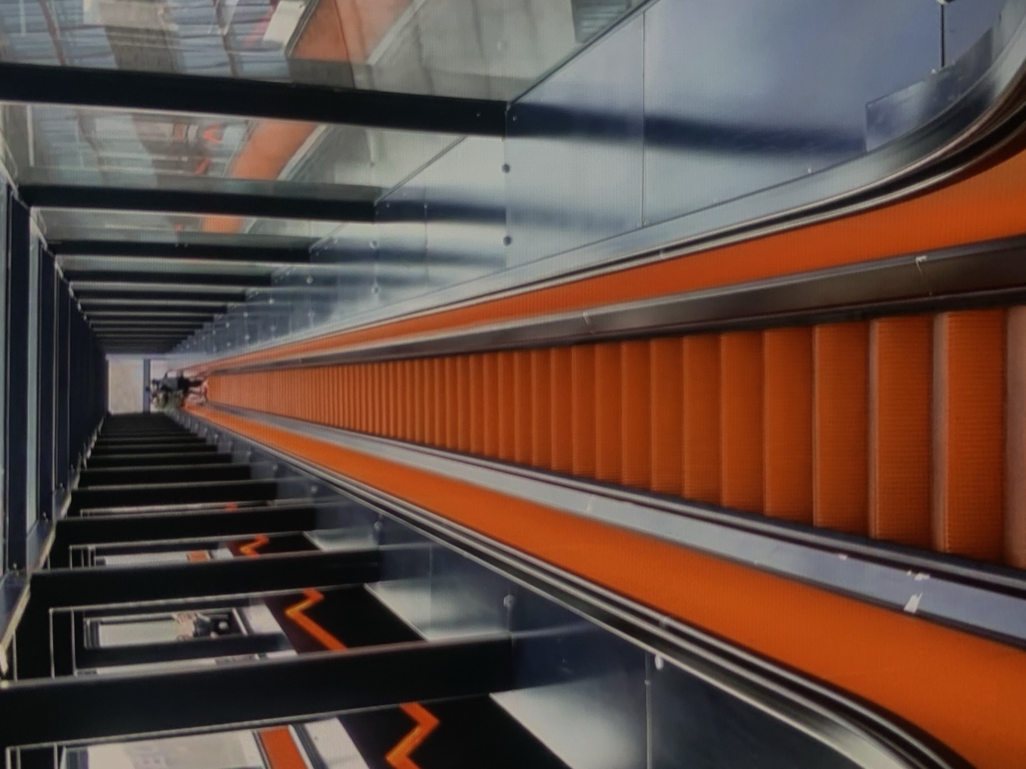 The image shows a long, orange escalator ascending within a covered walkway. The walkway is lined with black vertical pillars and large glass panels, offering glimpses of the surrounding environment which includes other escalators and architectural elements. Two people are visible at the top of the escalator, walking away from the camera. The scene suggests a modern, possibly public transport-related, infrastructure.