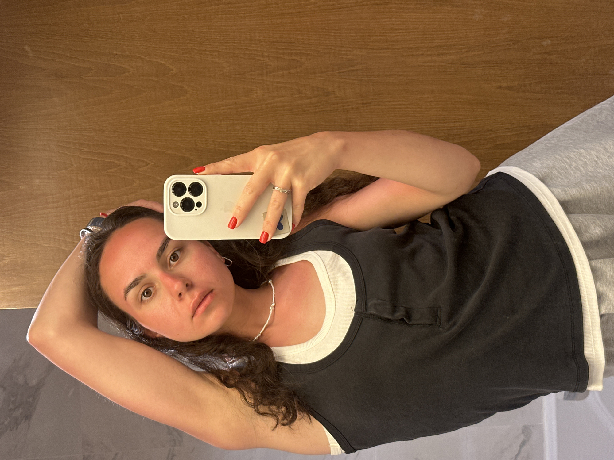 A woman is taking a selfie in what appears to be a bathroom. She is holding a white iPhone. She is wearing a black tank top over a white tank top, grey sweatpants, and a pearl necklace. Her nails are painted red. The background includes a wooden wall and a marble-tiled wall. No information suggests the location is in Pattaya City, Thailand.