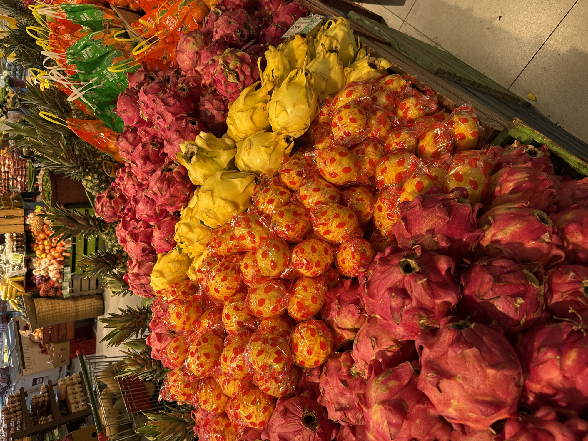 The image displays a vibrant, abundant produce section, likely within a grocery store or market. The primary subjects are large quantities of fresh fruits arranged for sale. Prominently featured are piles of red dragon fruit (pitaya), bright yellow dragon fruit, and individually wrapped, round, yellow-red patterned fruits, possibly Asian pears or quinces, encased in clear plastic. Rows of pineapples with their green tops are also visible, along with oranges or tangerines packaged in green and orange mesh nets. In the background, other produce like onions, and various packaged goods on metal shelving and in baskets, can be seen.
The scene depicts a static display of fruits, implying a shopping environment without any active human presence. The fruits are neatly organized, with distinct sections for each type and color, creating a visually appealing arrangement. The lighting is artificial, characteristic of indoor retail spaces, casting some soft shadows.
Notable visible text includes "VALENCIA MANDARIN" on the colorful mesh bags. The clear plastic wrapping on the round fruits shows partial text such as "Long-Fu... Fresh... Fruit" and "Premium Quality". A price tag in the yellow dragon fruit section partially reads "900.5", and a box in the distant background has "INDULGE" printed on it. The floor appears to be tiled.