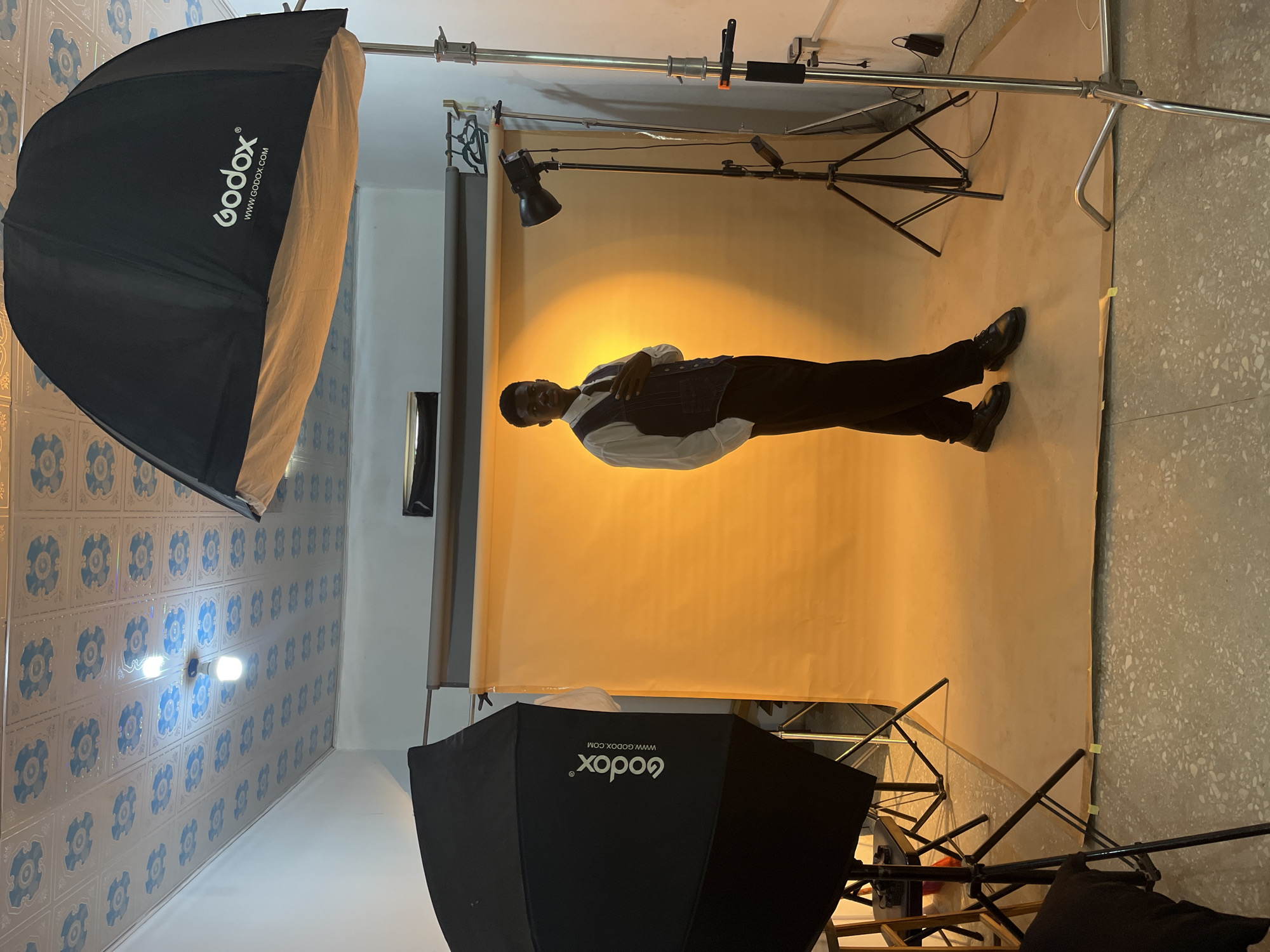 The image depicts a man posing in a photography studio in Gura Top, Nigeria. He's wearing black trousers, a white shirt, and a dark-colored waistcoat. He is positioned in front of a large, tan backdrop illuminated by studio lighting. The room includes studio equipment, notably two large softboxes with the brand "Godox" visible on them. One softbox is placed on the left side, and the other is partially visible at the bottom. Light stands support the equipment. The room's walls are decorated with blue and white patterned tiles. The floor is made of terrazzo.