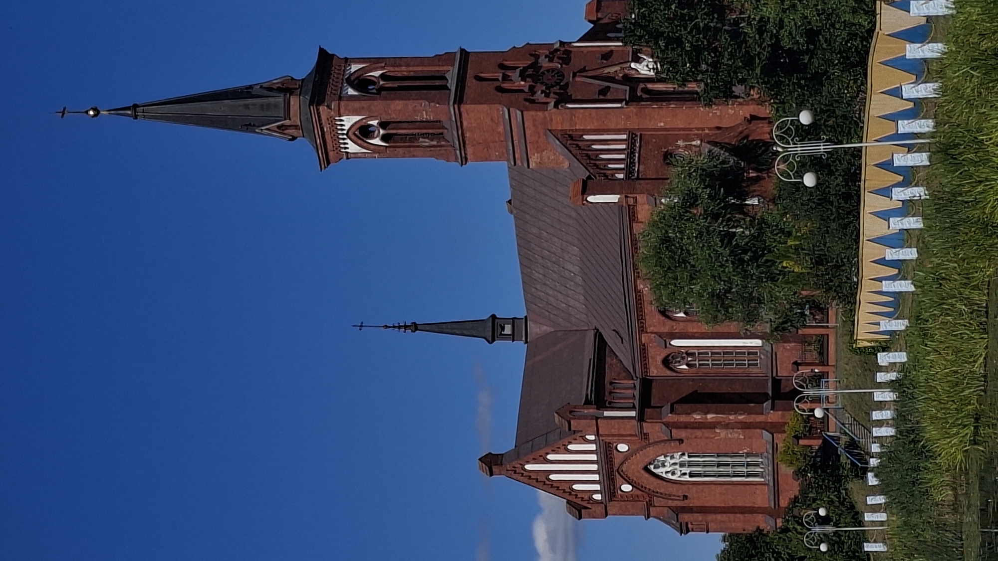 A majestic red brick church with a tall, dark spire stands prominently against a clear blue sky in Pastavy, Belarus. The church features gothic architectural elements, including arched windows with white detailing and a smaller spire atop the main structure. The foreground is dominated by lush green vegetation and reeds, bordering what appears to be a body of water. A decorative fence with a yellow and blue zigzag pattern runs along the edge of the water, interspersed with white posts. Several lampposts with white globes are visible, suggesting a public space or park. Trees and bushes are scattered around the church, partially obscuring its lower sections. The overall scene depicts a serene and picturesque view of a religious landmark.