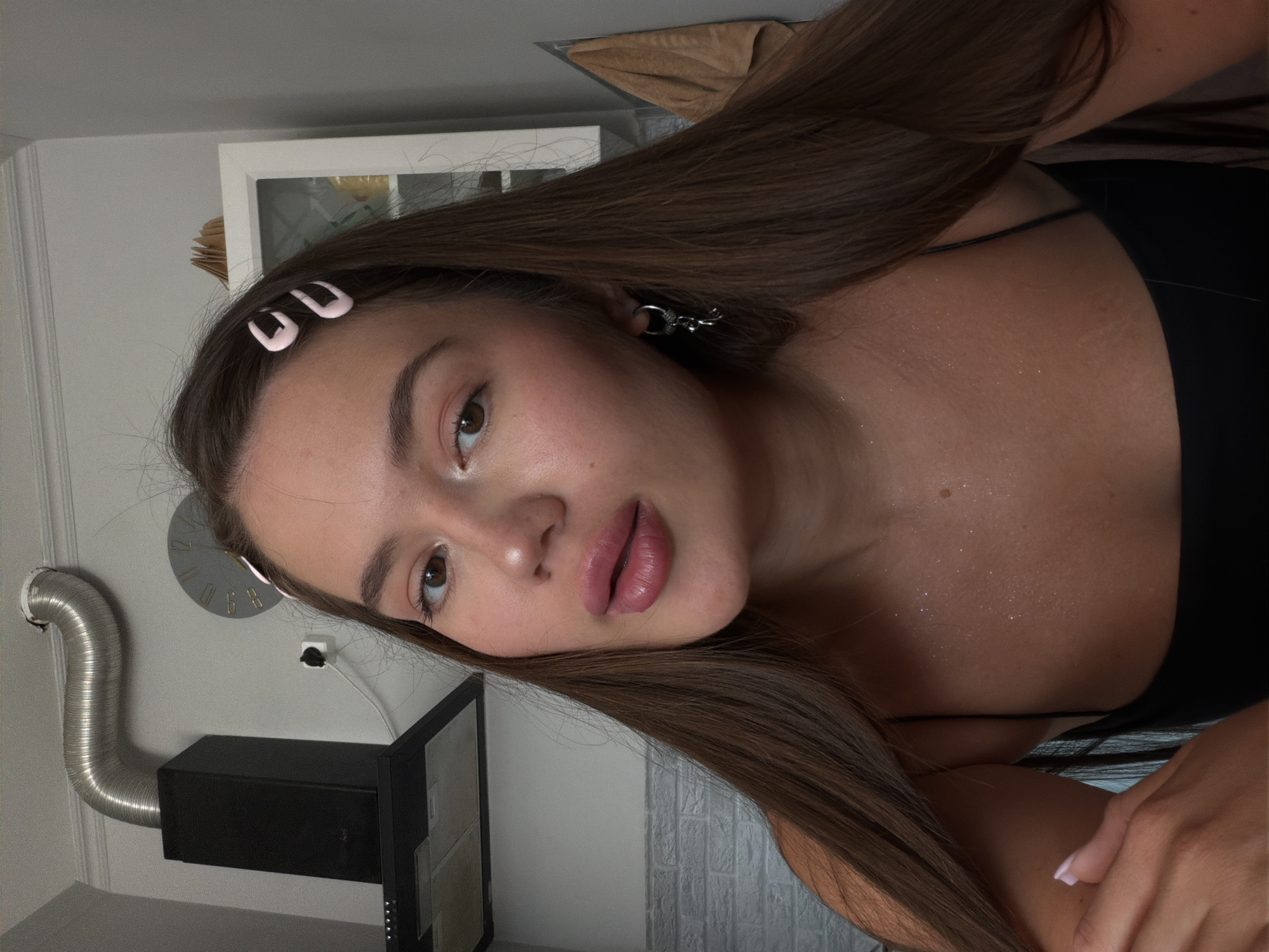 A young woman is captured in a close-up, selfie-style shot, likely in a kitchen setting. She has long, dark brown hair adorned with two light pink hair clips on the right side of her head. Her makeup includes a notable application of lip gloss, accentuating her full lips. She is wearing a black, spaghetti-strap top, and her décolletage is visible, enhanced with what appears to be body glitter, creating a shimmering effect. A silver dangle earring is visible on her left ear. In the background, a black range hood with a corrugated metal vent pipe is mounted on a light grey wall. To the right of the hood, a round clock with a minimalist design is present. Further to the right, a white cabinet with glass doors is partially visible, and a brown towel hangs on the wall. The overall ambiance suggests an indoor, domestic environment. The mention of Sirius, Russia, in the prompt is not supported by visual cues within the image.