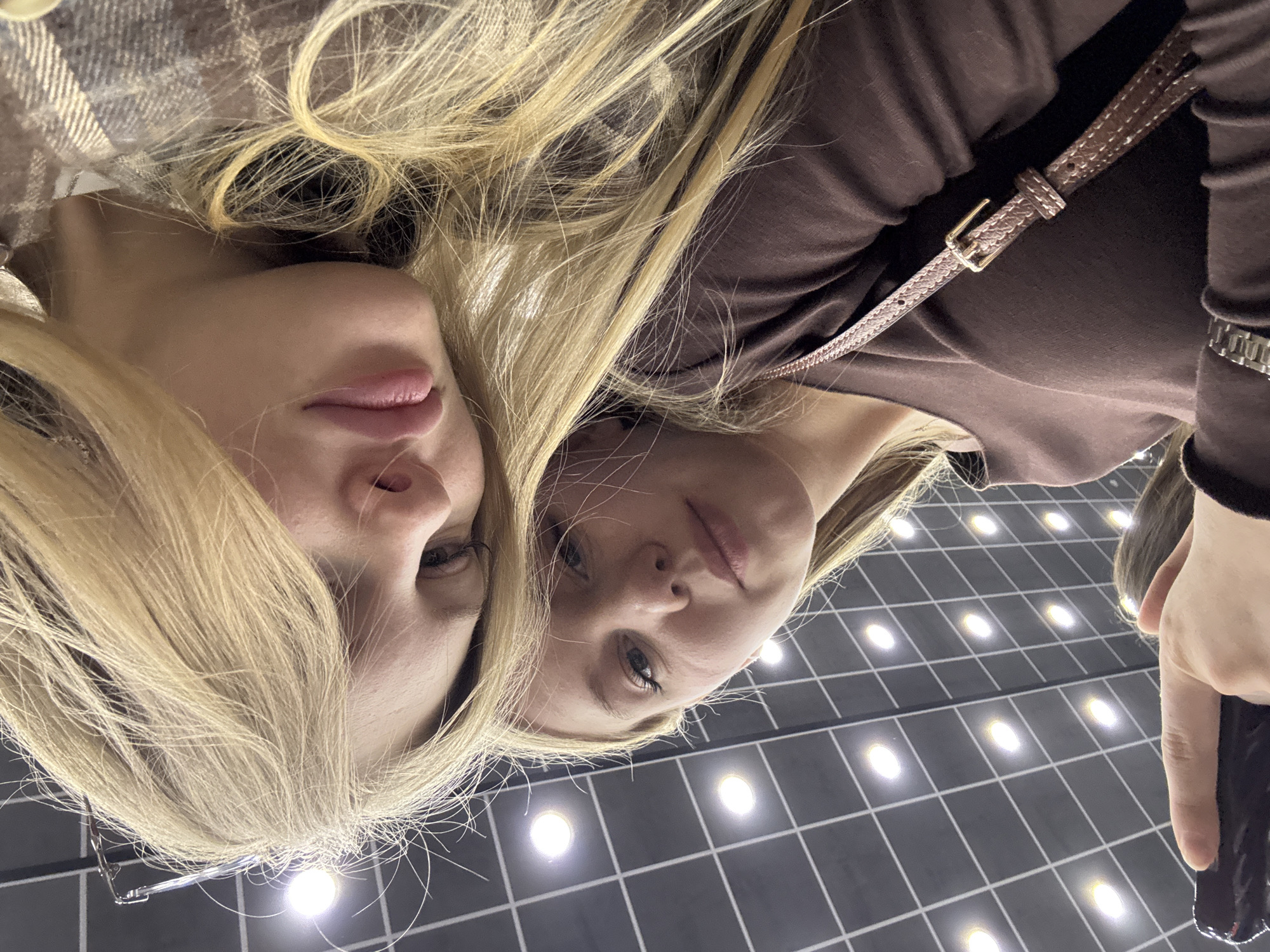 Two blonde-haired women are captured in a low-angle shot, possibly a selfie looking downwards or a reflection. The woman on the left wears a plaid or checkered garment and has glasses hanging near her ear. The woman on the right is dressed in a plain brown long-sleeved top, with a brown, textured strap featuring a gold buckle crossing her chest, and a silver-toned watch on her left wrist. Both women have neutral to slightly serious expressions, with their gazes directed towards the camera. Their light-colored hair flows across the frame, mingling between them.
The background is a dark, gridded surface, likely a floor or ceiling, interspersed with numerous evenly spaced, circular bright lights that illuminate the scene. This unique pattern suggests a modern architectural setting, such as a contemporary public space, shopping mall, or an art installation in Perm, Russia. The lighting is entirely artificial, indicating an indoor environment. No specific activity beyond posing is evident, and no visible text is present.