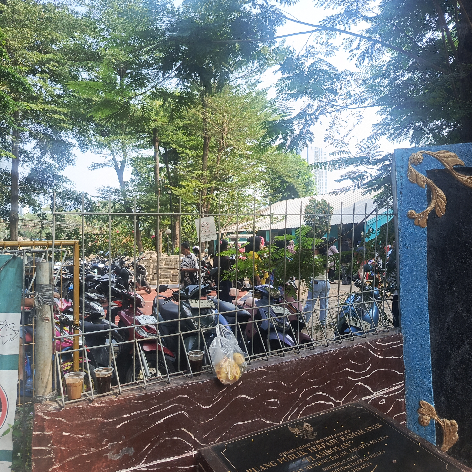 In South Jakarta, Indonesia, a fenced-in area displays a collection of parked motorcycles under the shade of mature trees. Through the metal fence, a group of people are visible, some standing and others appearing to be walking. A building with a white roof is situated in the background, and beyond that, a modern high-rise building can be seen. A small sign with text and a graphic is attached to the fence. Two plastic cups, one containing a brown liquid, rest on the fence's top rail. A plastic bag filled with yellow, circular items hangs from the fence, positioned above a low, textured wall. A plaque with inscribed text and an emblem is mounted on the wall. The overall scene suggests a public space with a parking area and surrounding natural and urban elements.