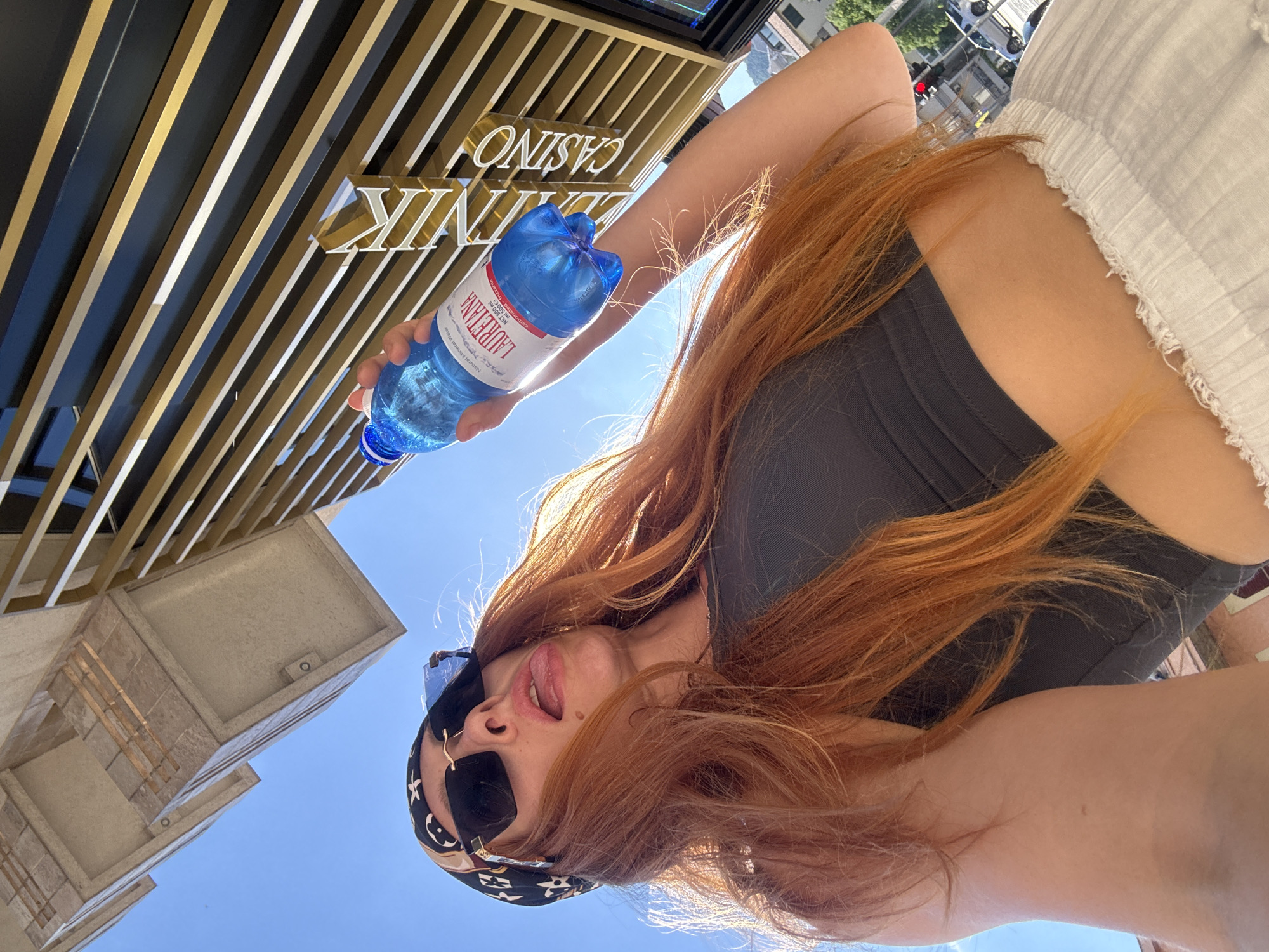 In Budva, Montenegro, a woman with long, reddish-orange hair and sunglasses is taking a selfie outdoors, holding a bottle of Lauretana natural mineral water. She is wearing a black crop top and white, textured pants. Her tongue is sticking out, and she is looking upwards towards a building with a golden-slatted facade and signage that appears to read "Casino". The sky is clear and blue. The image is taken from a low angle.