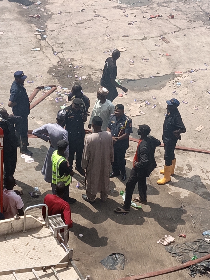This image depicts an outdoor scene in Fagge C, Nigeria, showing approximately 14 individuals gathered on a cracked, concrete surface under bright daylight. The ground is littered with small wrappers, plastic debris, and exhibits dark, wet-appearing patches. Several dark, coiled fire hoses are visible across the foreground and midground.
The group consists of both uniformed personnel and civilians. Six individuals wear dark blue or black uniforms, indicative of police or emergency services; some are equipped with caps, insignia, and holsters. One uniformed person stands out with bright yellow rubber boots, while another wears a yellow-green reflective safety vest over a dark uniform.
Civilian individuals are dressed in various attires: three wear light-colored traditional robes and caps, one wears a red t-shirt, one a light pink t-shirt, one a black long-sleeved top with dark trousers, and one a dark t-shirt. A tall individual in a black t-shirt is walking away from the main cluster towards the upper right.
The central group of individuals appears to be conversing or observing the scene. Shadows are long and distinct, cast by the bright sun. A metal structure, possibly part of a vehicle's ladder or platform, is visible in the lower-left foreground.