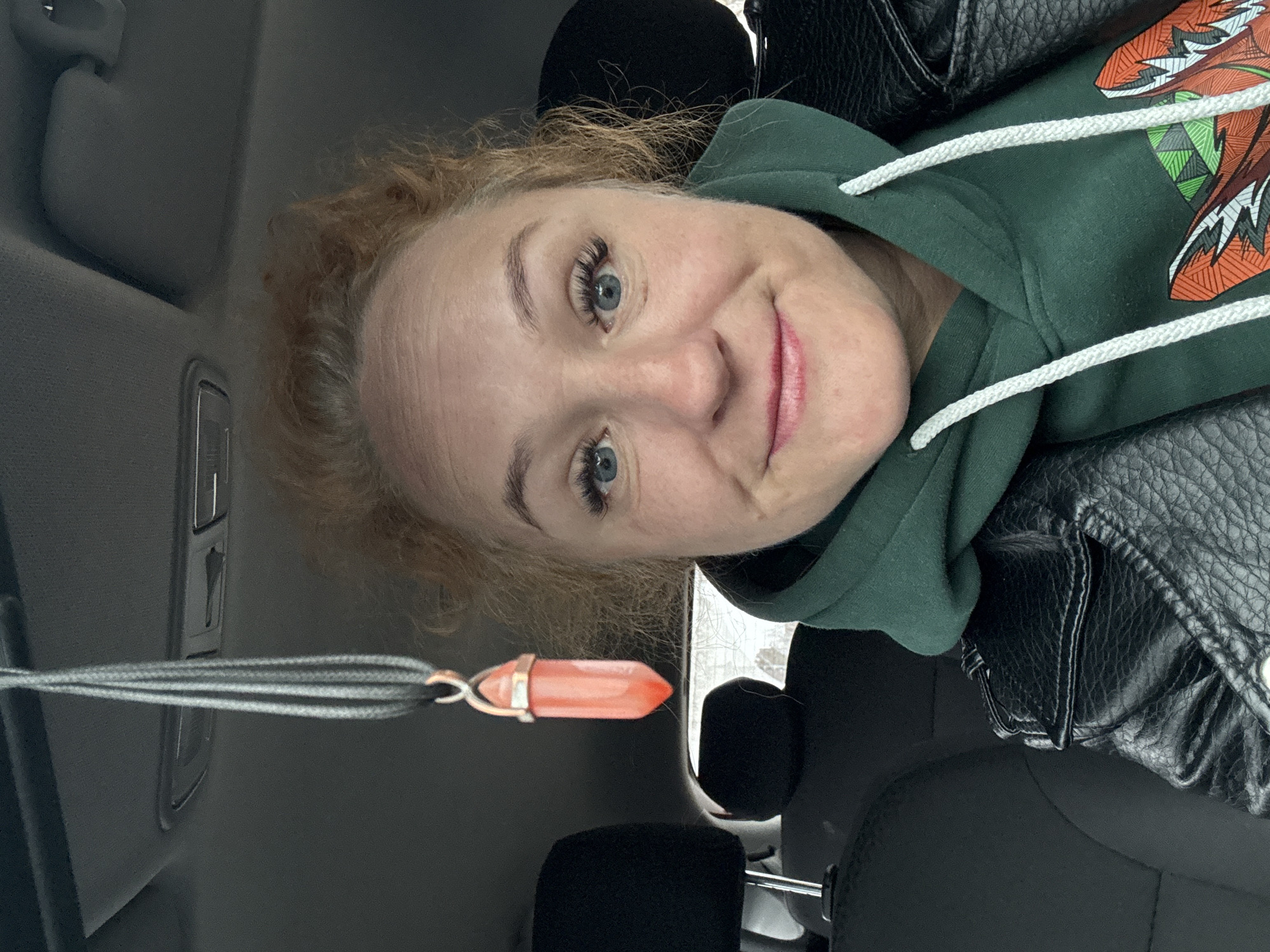 A woman with fair skin, blue eyes, and reddish-blonde curly hair is in the driver's seat of a car. She is wearing a green hooded sweatshirt with a design on the front and a black leather jacket. A grey necklace with a pink crystal pendant hangs in front of her. The car's interior is visible, including the dashboard and seats. The location is Chaikovsky, Russia.