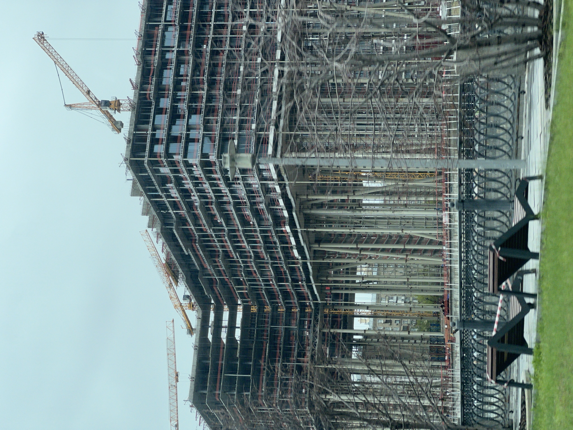 The image depicts a construction site in Moscow, Russia. A tall, unfinished skyscraper dominates the view, its steel frame exposed and several cranes attached to its upper levels. Visible through the steel frame is another building under construction. A wrought iron fence and bare tree branches are in the foreground and sidewalk with grass on the right side of the image. The sky is overcast.