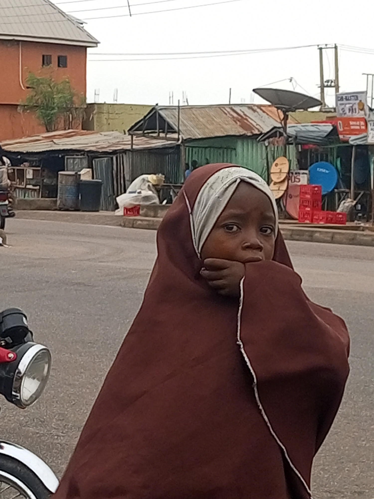 A young girl wearing a brown hijab and a white headscarf is the central focus of this image, her face partially obscured by her hand as she rests her chin on it. She is looking directly at the camera with a thoughtful expression. The girl is standing on a paved road, with a motorcycle partially visible in the foreground on the left side of the frame. In the background, there are several buildings with corrugated metal roofs, some appearing to be shops or stalls. Various objects are visible, including satellite dishes, stacked red crates, and what appear to be large barrels. The sky is overcast. The scene is consistent with a street view in Tumfure, Nigeria, likely a market area or roadside setting.