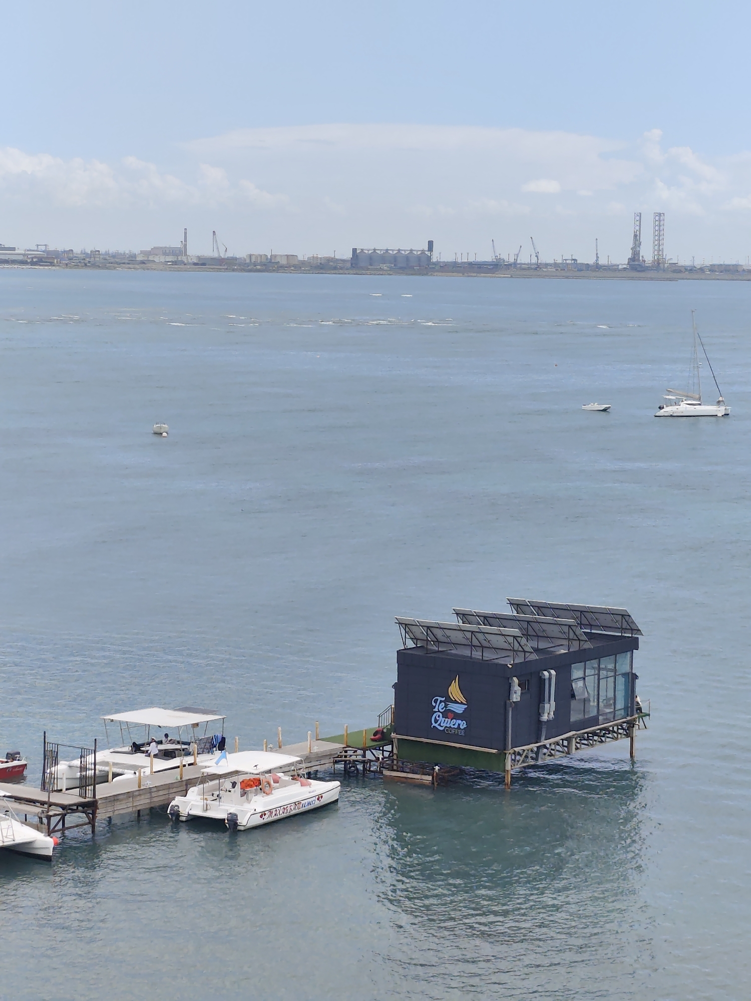 This media file depicts a waterfront scene in Aktau, Kazakhstan. In the foreground, a dark, modern floating structure with "Te Quiero Coffee" visible on its side is anchored to a wooden dock. The structure has large windows and solar panels on its roof. Several boats, including speedboats and a larger catamaran with a canopy, are moored to the dock.
In the mid-ground, the water is calm, with a few smaller boats and buoys visible. In the background, a distant industrial shoreline stretches across the horizon. Silos, cranes, and tall structures suggest a port or industrial area. The sky is partly cloudy, indicating fair weather. The time of day appears to be daytime, possibly late morning or early afternoon, given the lighting. The overall mood is peaceful and serene, with a focus on leisure and industry coexisting.