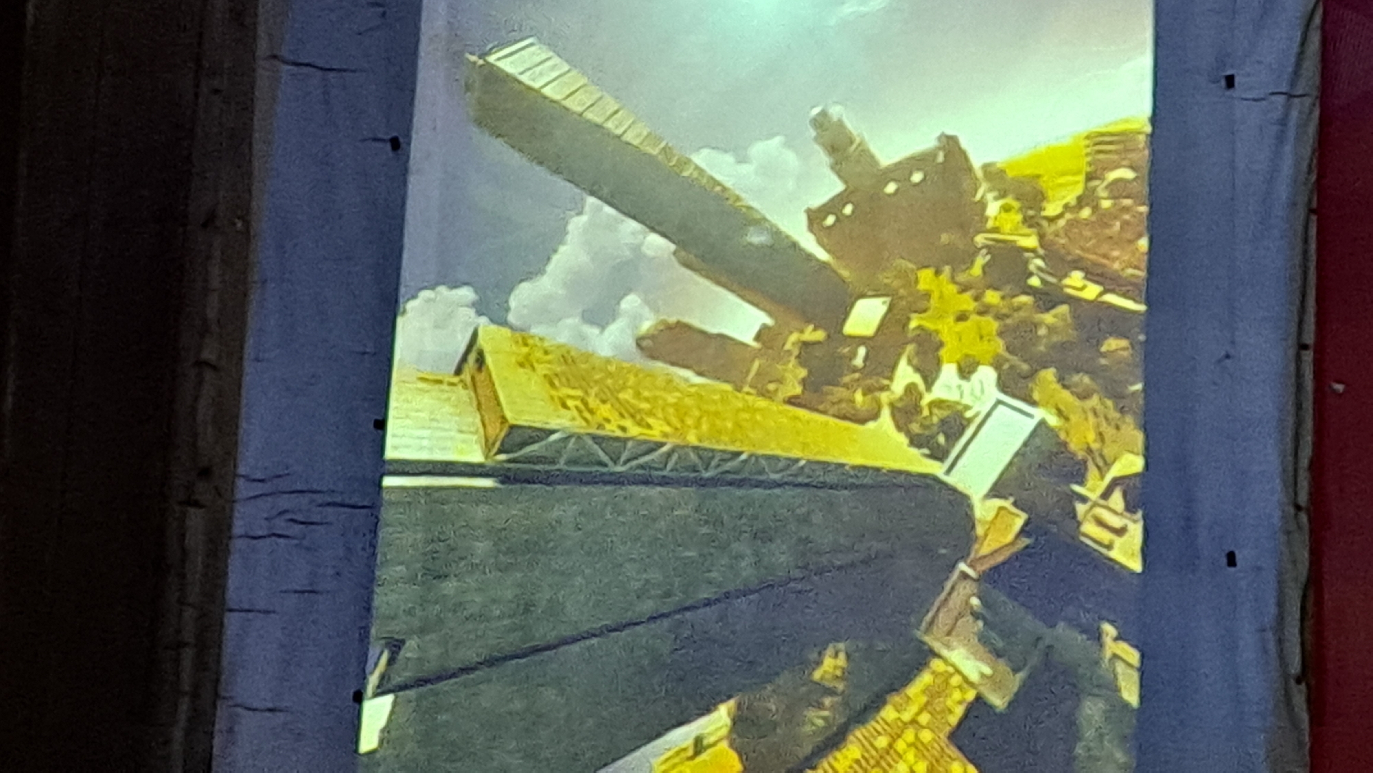 The image displays a projection of what appears to be an artistic rendering of skyscrapers. The architectural style suggests a futuristic or stylized representation rather than a realistic depiction of a specific city. The skyscrapers are tall and angular, with a metallic or glass-like sheen. Some are depicted in shades of gold and dark gray, while others have a more abstract, textured appearance. Clouds are visible in the background, suggesting an outdoor setting or a sky element within the artwork. The overall impression is one of urban grandeur and possibly a sense of height and vastness. There is no indication in the visual information provided that this scene is located in Payagaboh, Indonesia, or any other specific geographical location. The projection is displayed on a dark surface, possibly a screen or wall, with visible texture and some minor imperfections in the projection area, suggesting it's a real-world viewing of a digital or projected artwork.