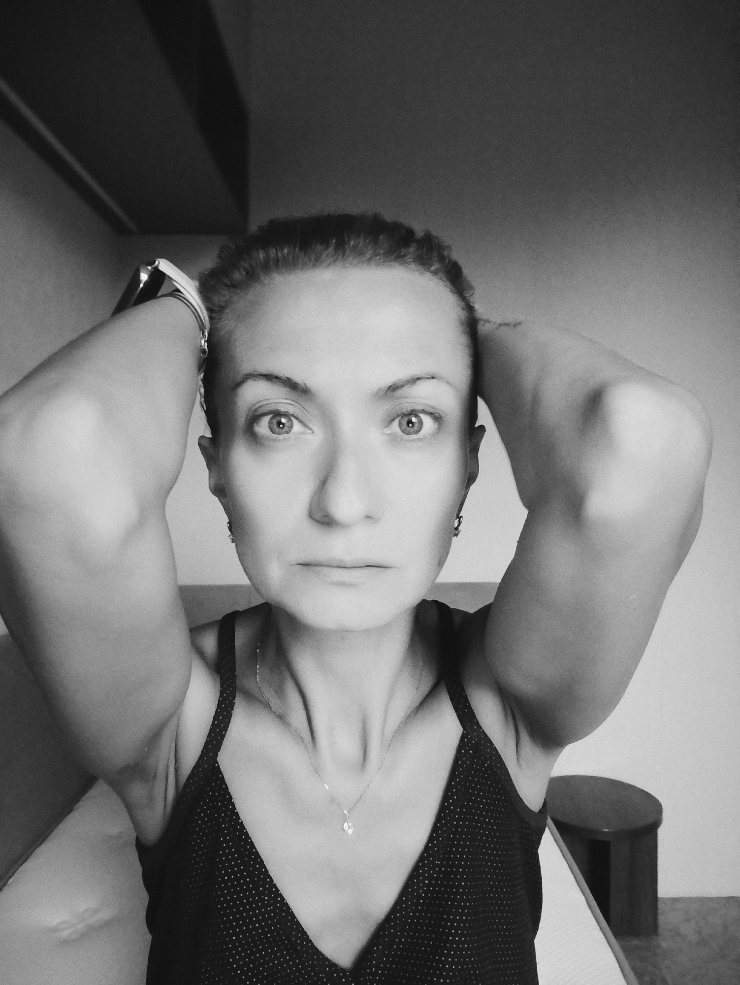 A black and white, close-up, frontal shot shows a woman from the chest up, with her arms raised and hands on the back of her head. She has light skin, dark hair pulled back, and is wearing a dark, polka-dot tank top and a delicate necklace with a small pendant. Her eyes are wide open and looking directly at the camera. The background is a plain wall, with what appears to be ceiling beams casting shadows. No specific location in Moscow, Russia, is identifiable.