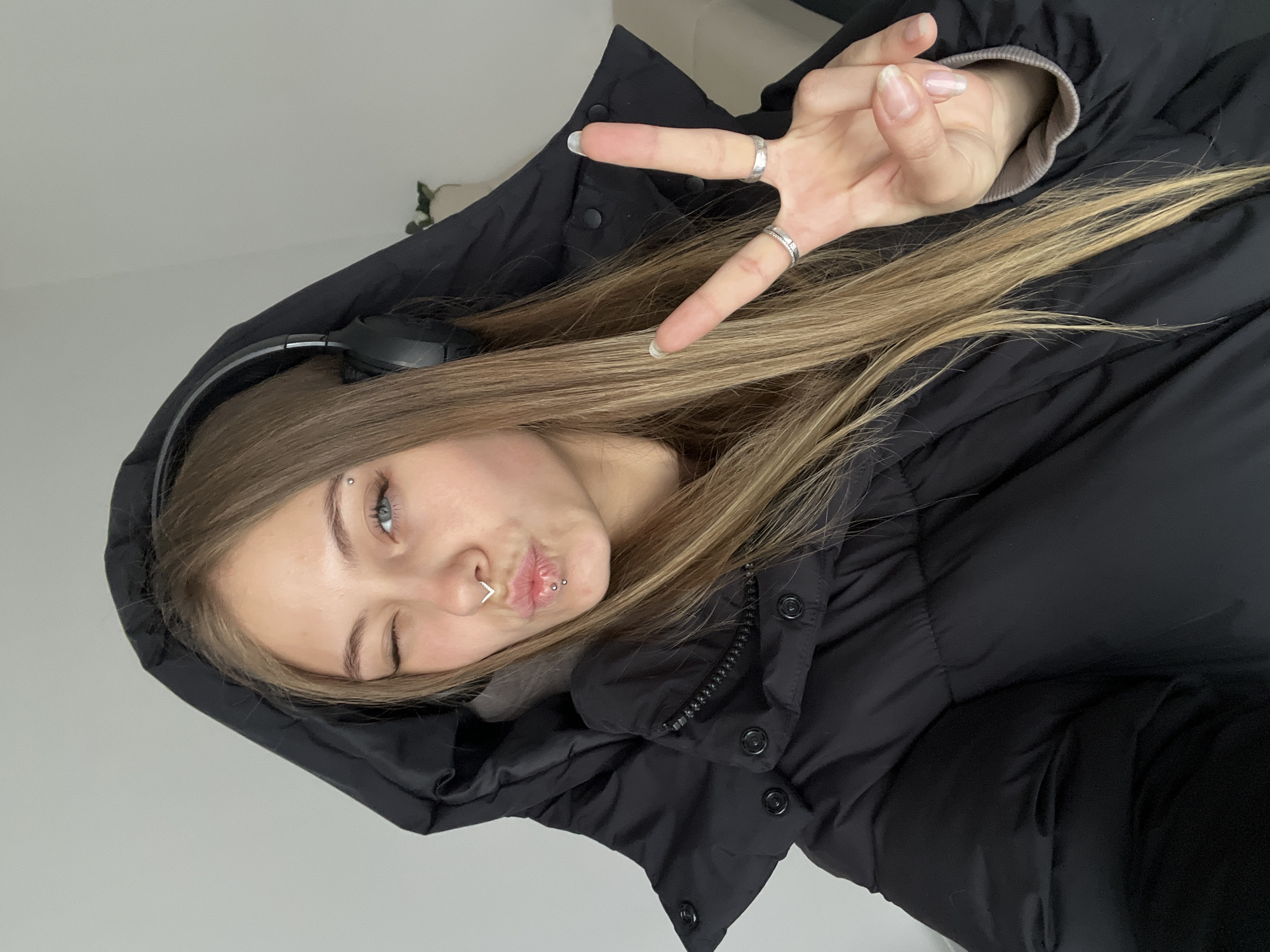 A woman in a black hooded puffer jacket is making a peace sign with her right hand. She is winking her left eye. She has light brown hair and is wearing headphones. She has multiple piercings on her face including a nose ring, eyebrow piercing, and lip piercings. The background is a plain white wall. There is no indication of the city of Фоки, Russia in the image.