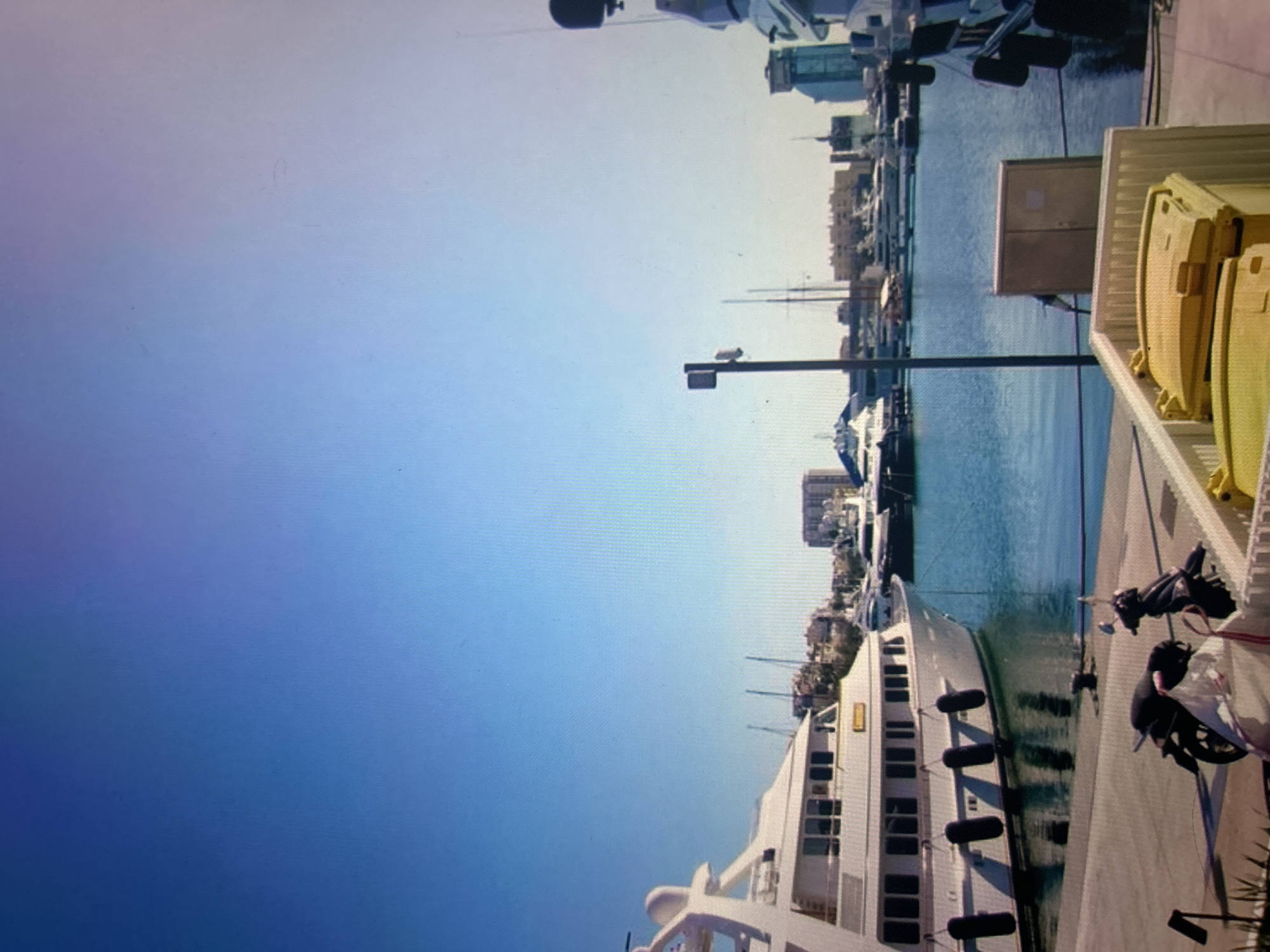 The image captures a vibrant marina scene on a clear, sunny day, with the perspective rotated 90 degrees counter-clockwise. When viewed correctly, a bright blue sky dominates the upper portion, transitioning from a lighter hue at the horizon to a deeper blue overhead.
In the middle ground, the serene blue water of the marina stretches horizontally, reflecting the light. Numerous boats and yachts are docked along the water's edge, with several masts visible against the sky. A prominent, large white yacht with multiple windows and black fenders along its side occupies a significant part of the lower-left and central frame, moored alongside a paved or concrete dock.
The dock, visible in the lower right, features several items, including a tall black pole (possibly a light or camera pole) and large yellow containers, which appear to be storage bins or safety equipment. Further along the dock, next to the white yacht, there's a dark-colored object resembling a scooter or bicycle. In the background, across the marina, a cityscape or town is visible with various buildings, suggesting an urban or semi-urban coastal setting.
The overall impression is one of leisure and calm, characteristic of a sunny day at a harbor. There are no people clearly visible, and the boats appear to be static, indicating a quiet moment in the marina. No discernible text is visible on any objects or buildings in the image.