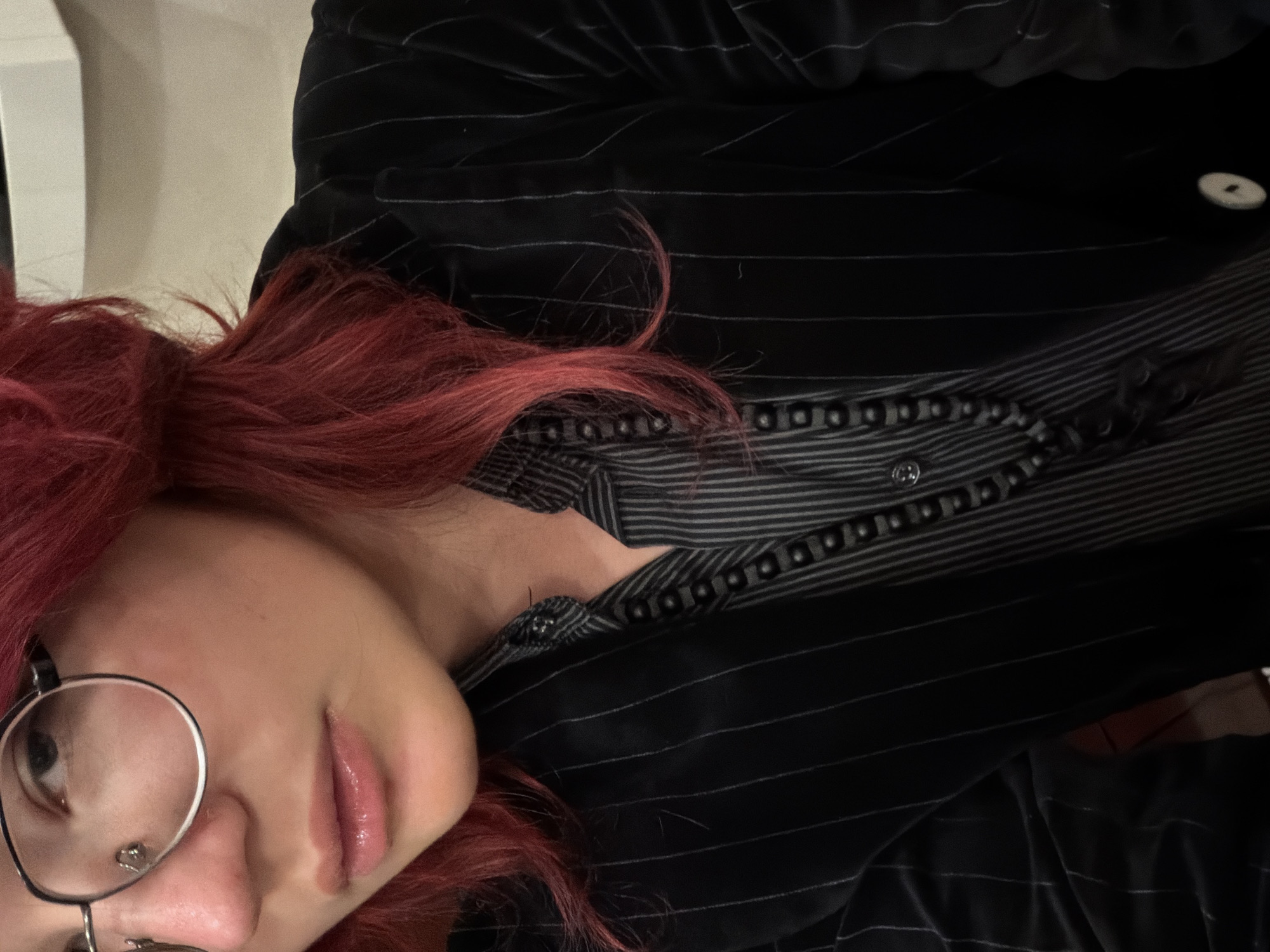 A close-up, chest-up, medium shot shows a person with bright red hair, wearing glasses with round frames, a striped collared shirt, and a pinstriped jacket. The person is looking slightly to the left of the camera, their lips are slightly parted and glossy. They are wearing a dark, beaded necklace. The background is out of focus and appears to be indoors, possibly a dimly lit room or restaurant. There is no information in the image to suggest a location in Chelyabinsk, Russia.