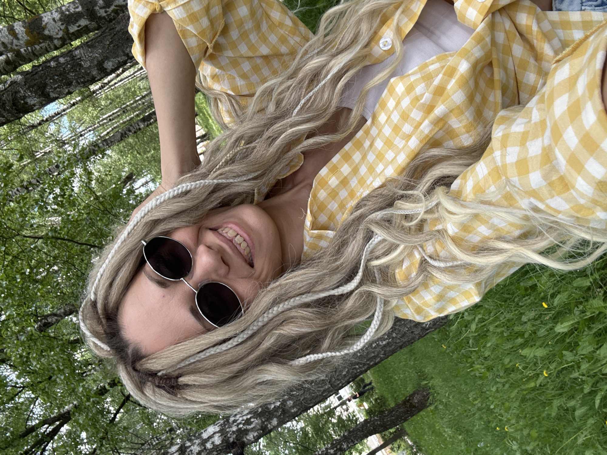 A woman with braided blonde hair and round sunglasses is lying on her back in a grassy area with trees, possibly in the city of Шебунино, Russia. She is wearing a yellow and white checkered shirt over a white top. The image is angled, capturing the woman's smiling face, the details of her hairstyle, clothing, and the surrounding greenery. The presence of trees suggests the location might be a park or wooded area.