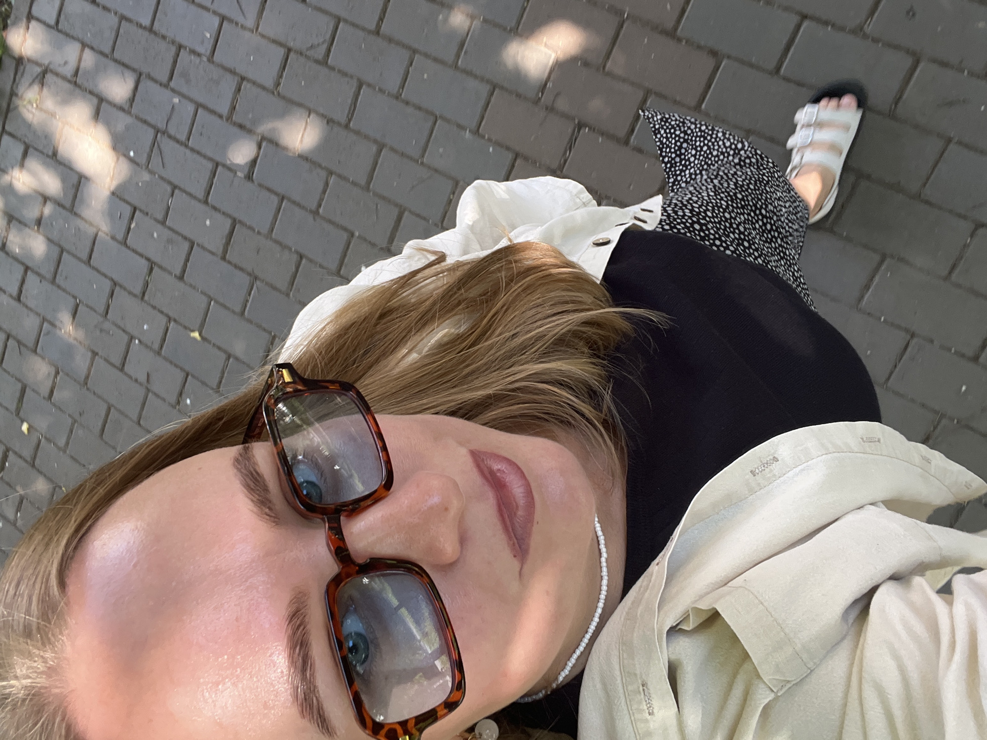 This media file shows a close-up, overhead, first-person perspective of a person outdoors. The subject is wearing a black and white polka dot dress, a white button-up shirt, and white sandals. They are also wearing tortoiseshell glasses and a beaded necklace. Their blonde hair is visible. The background is a gray brick pathway with dappled sunlight creating shadows. The person appears to be walking, as indicated by the angle of their foot and the context of the pathway. The lighting suggests it is daytime, likely sunny. The setting appears to be a sidewalk or pedestrian area.