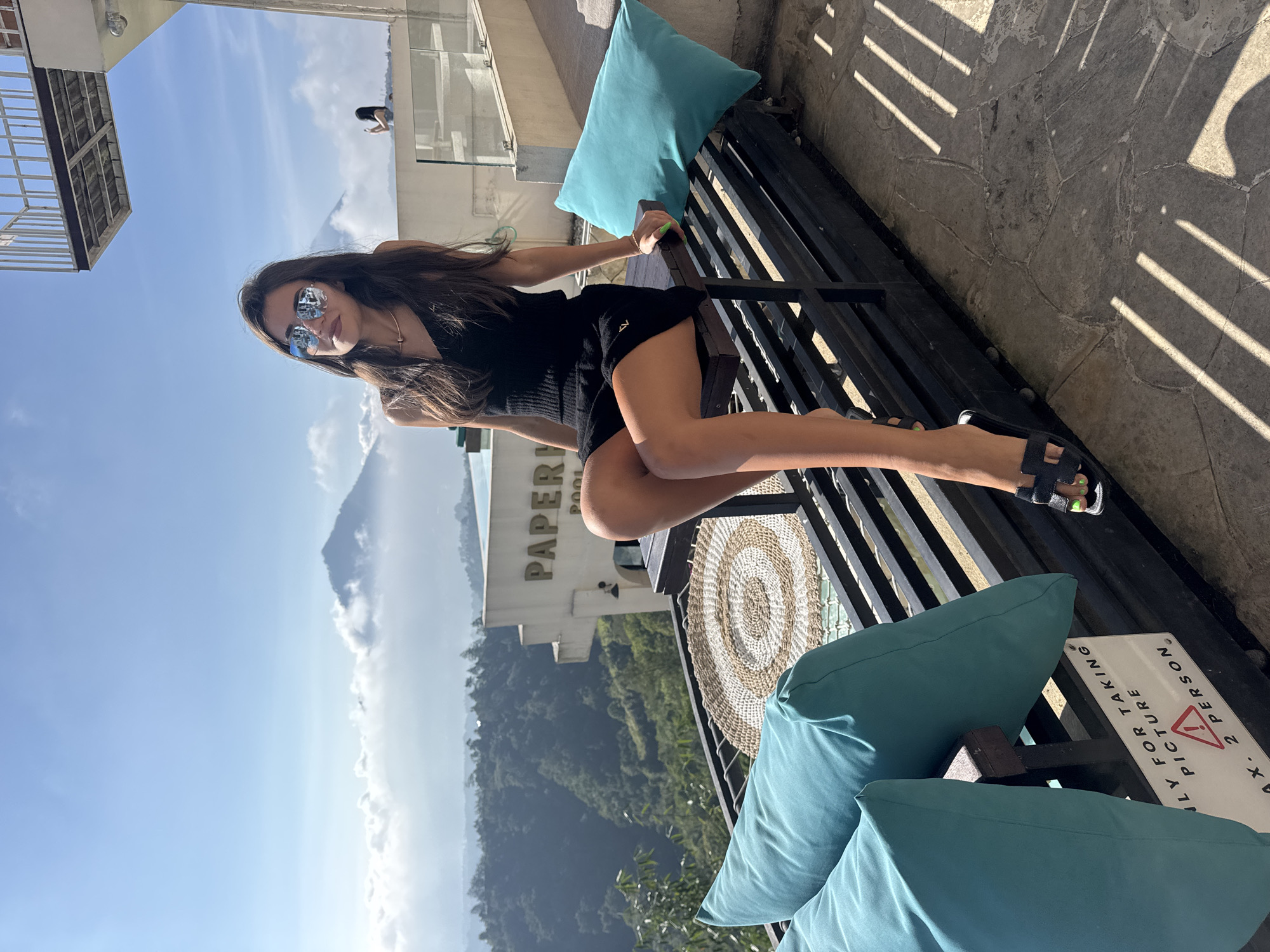 A woman is sitting on a bench on a balcony overlooking a valley in Kintamani, Indonesia. She is wearing a black tank top, a black skirt, and black sandals. She has long, dark hair and is wearing sunglasses. She is holding a phone in her right hand. The balcony has a railing and a few pillows. In the background, there is a building with the word "PAPER" on it. There are also mountains and clouds in the distance. The sun is shining and the sky is blue.