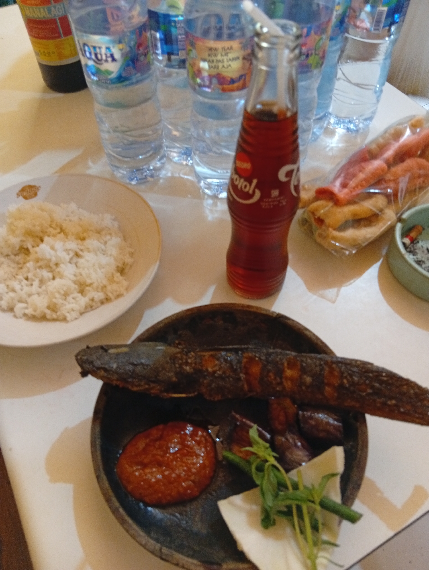 The image shows a close-up, overhead view of a meal laid out on a table, likely in a casual dining setting. The meal consists of white rice served on a white plate with a gold rim, a deep-fried fish, a small dollop of red chili paste, and some green vegetables that appear to be long beans and herbs. Beside the rice is a small bottle of what appears to be a dark red soda, possibly a local brand. In the background, several large plastic bottles of water are visible, along with a packet of snacks that look like puffed crackers. To the right, a small ceramic ashtray contains cigarette butts and ash. The overall lighting suggests an indoor setting, possibly a restaurant or home. The meal presentation is informal, and the presence of water bottles and snacks implies a relaxed dining experience. The location is stated to be in the city of Ubud District, Indonesia.