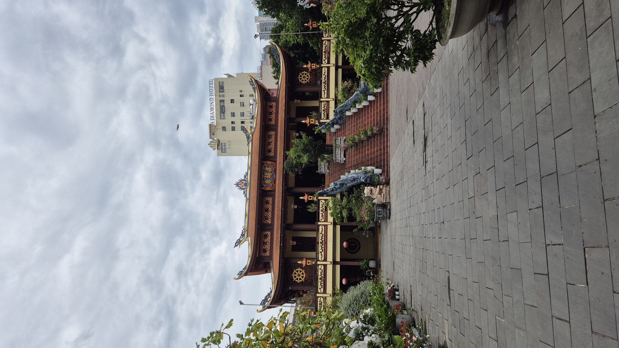 The image captures a view of a Buddhist temple with traditional architecture, featuring a multi-tiered, ornate roof with upturned eaves, painted in shades of red and gold. The temple is approached by a wide, paved walkway leading to a grand staircase flanked by dragon sculptures and ornate railings. Numerous potted plants and decorative elements, including stone statues, are placed around the temple entrance and along the walkway. In the background, a modern, multi-story building with the signage "HOA DIAMOND HOTEL" is visible, suggesting the temple is located in a more urban setting within the city of Đà Nẵng, Vietnam. The sky is overcast, contributing to a muted light. The scene is devoid of people.