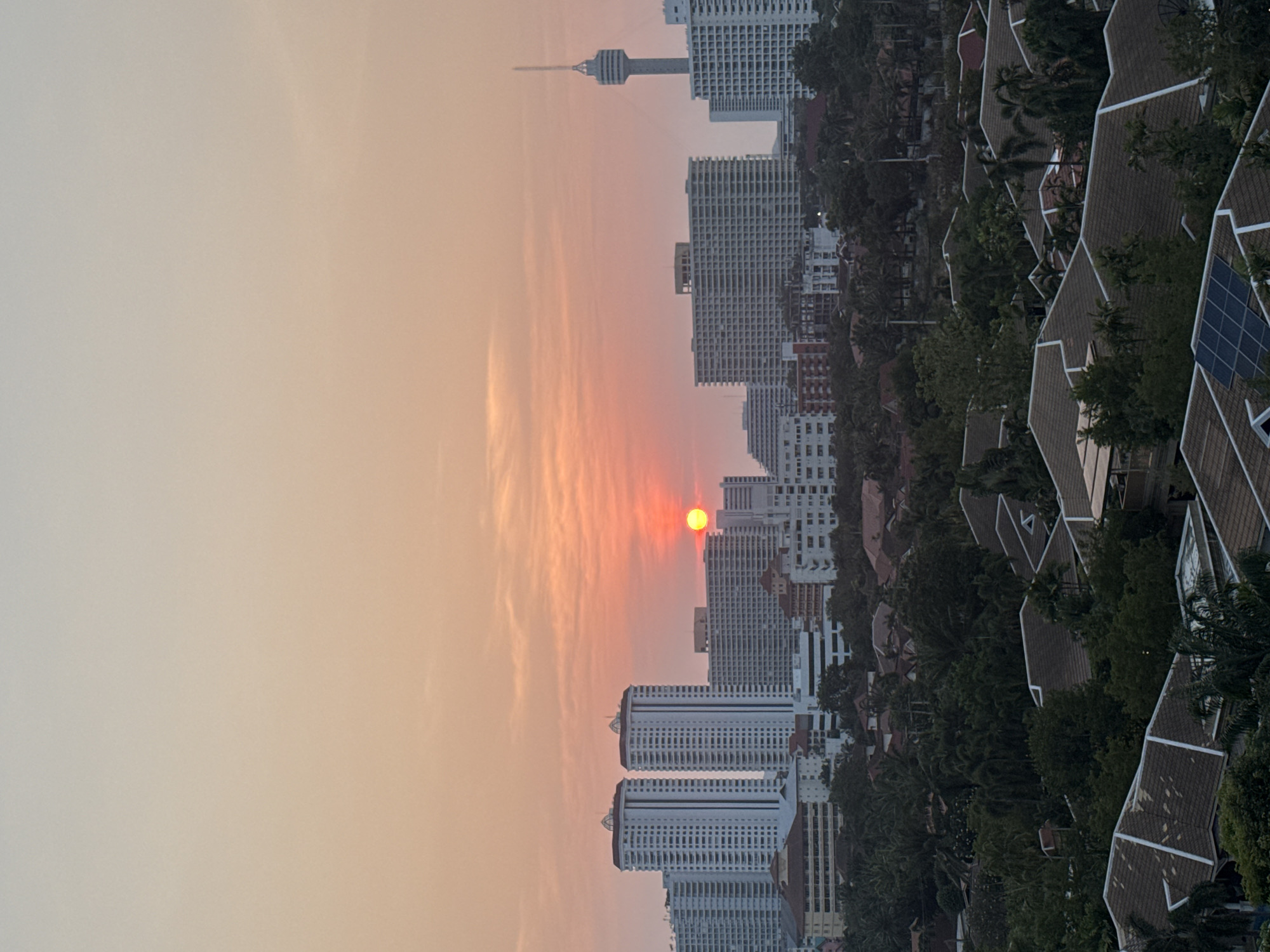 The image depicts a skyline at sunset in Pattaya City, Thailand. A large orange sun is visible amidst the high-rise buildings and condominiums, contributing to the overall reddish-orange hue in the sky. There is a communications tower visible in the background. The right side of the frame showcases rooftops of various buildings interspersed with trees. The photograph appears to have been rotated 90 degrees counterclockwise.