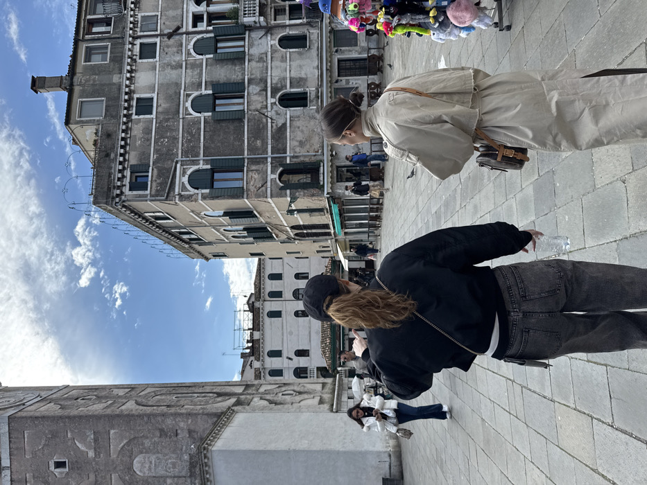 The image displays an outdoor urban scene in Campo San Giacomo di Rialto, Venice, Italy, under a sky with scattered white clouds. The ground is paved with rectangular grey stone blocks.
Two females are centrally positioned in the foreground, viewed from behind, walking forward. The female on the left wears a black baseball cap, a black bomber jacket, a white shirt, and dark grey, relaxed-fit jeans. Her light brown hair is tied in a ponytail, and she holds a clear plastic water bottle in her right hand. A thin strap, likely from a small bag, crosses her back. The female on the right wears a long, light beige trench coat and carries a small brown shoulder bag with a prominent buckle on her right side. Her dark hair is styled in a bun.
To the left of the subjects, a tall, weathered grey stone bell tower, identifiable as the Campanile di San Giacomo di Rialto, rises prominently. It features arched openings and geometric patterns on its facade. A portion of a smaller, white building is visible at its base.
To the right of the subjects, a multi-story building with a heavily weathered, grey facade displays numerous arched windows, many fitted with dark green closed shutters. The ground floor contains storefronts with green awnings. Partially visible on the far right is a street vendor's stall displaying colorful plush items.
In the mid-ground, other pedestrians are visible walking or standing. Further down a passageway, additional Venetian buildings with white facades and green-shuttered windows are present. Several pigeons are scattered on the plaza ground. The scene is illuminated by natural daylight.