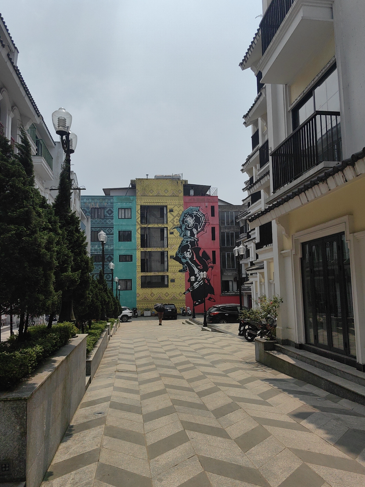 The scene depicts a pedestrian street in Sa Pa, Vietnam, under a bright, hazy sky. The walkway is paved with alternating light and dark gray rectangular tiles forming a prominent chevron pattern that extends into the background.
On the left side, a low, light gray stone retaining wall runs parallel to the path, behind which a line of tall, slender evergreen trees is planted. Interspersed within the tree line are decorative streetlights with clear, ribbed glass globes atop dark poles. Beyond these trees, a series of light-colored, multi-story buildings with dark tiled roofs and green balcony railings are visible.
The right side of the street features similar light-colored, multi-story buildings, with the structure closest to the foreground exhibiting a light yellow facade and a large dark-framed glass entrance. A black scooter or motorcycle is parked angled onto the sidewalk on this side.
At the far end of the street, a prominent multi-story building is the central focus. This building is vertically segmented into distinct colored sections: teal on the left, yellow in the center, and red on the right. Both the teal and yellow sections feature subtle light-colored geometric patterns. A large mural, primarily black with light blue accents, is painted across the yellow and red sections, depicting two stylized figures in traditional attire, with the upper figure holding an open umbrella.
A single individual is observed in the mid-ground, walking away from the viewer on the paved street, holding a dark open umbrella above their head. Several vehicles, including a white SUV and a black sedan, are parked near the base of the multi-colored mural building in the background.