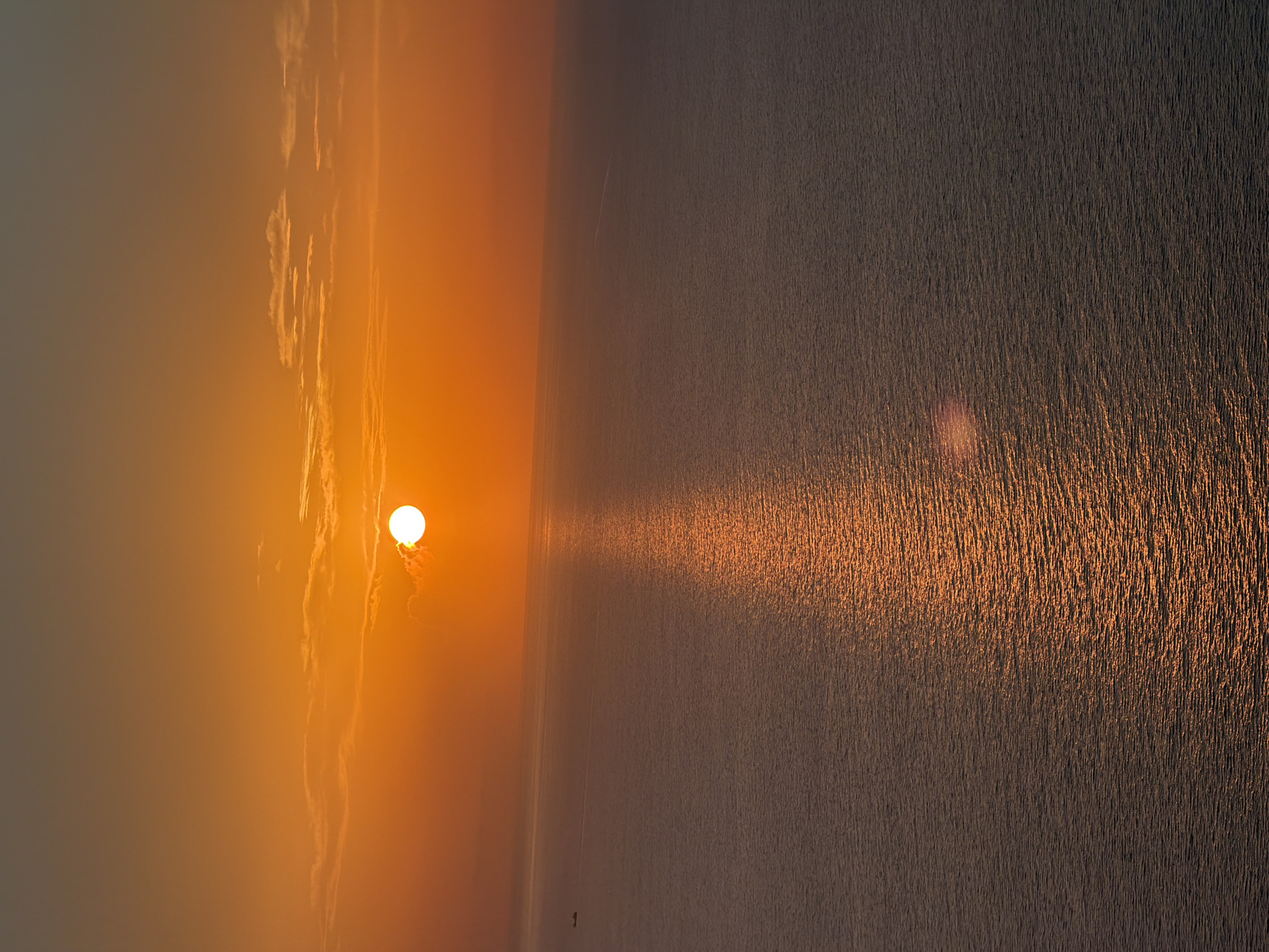 This is an aerial, full-frame, vertical shot of a sunset over the Mediterranean Sea, likely taken from a high vantage point in Sant Antoni de Portmany, Spain. The sun, a bright white disc partially obscured by wispy clouds, is positioned in the upper third of the frame, casting a shimmering golden path across the water. The sky transitions from a deep orange hue near the horizon to a muted gray at the top of the image. The sea, a deep blue-gray, is rippled with small waves, reflecting the warm light of the setting sun. A faint, dark shape resembling a boat or small vessel is visible on the left side of the image, moving across the water. The overall scene is one of serene beauty, capturing the tranquil ambiance of a Mediterranean sunset.