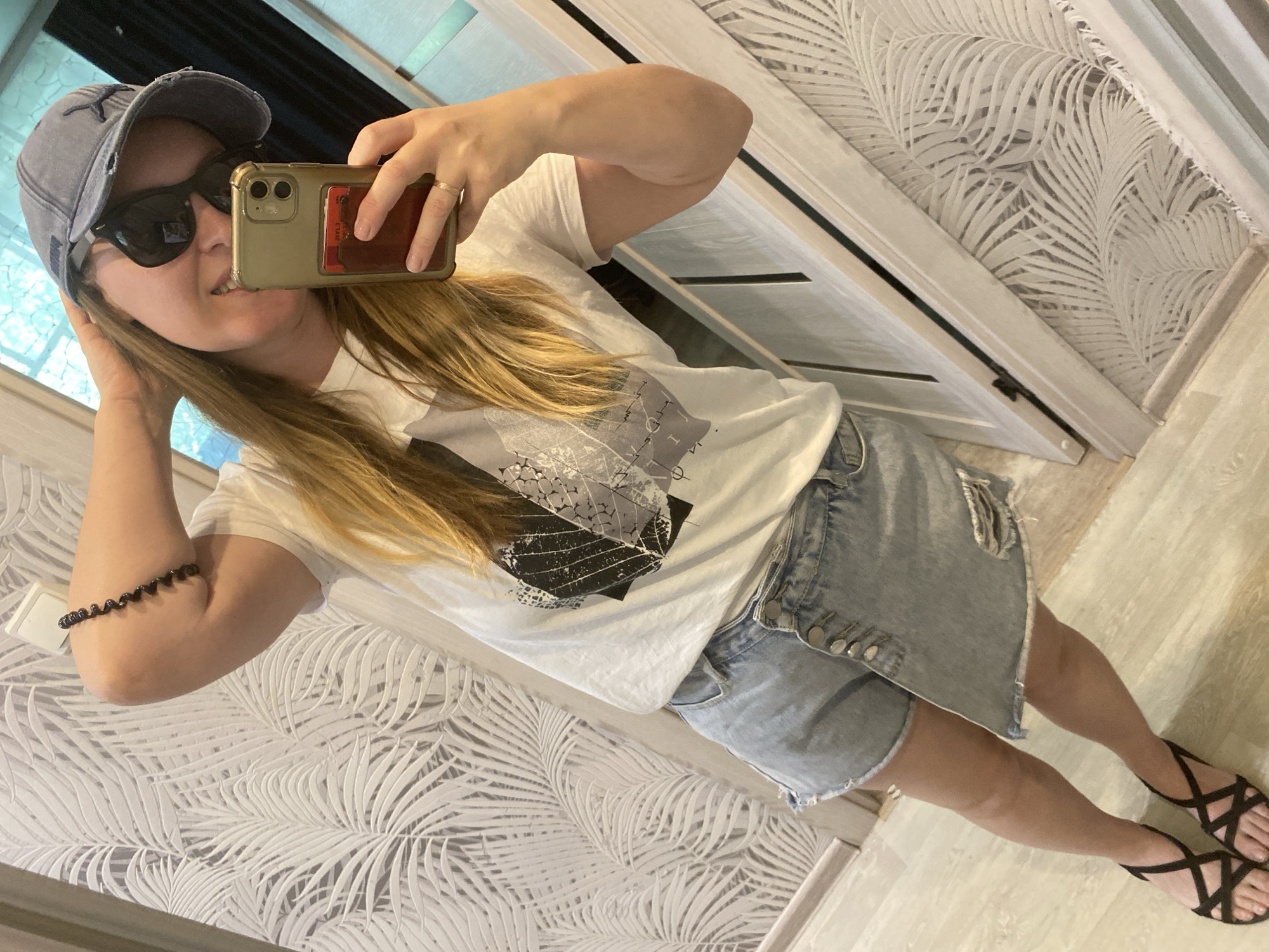 A woman is taking a selfie in what appears to be an interior space. She is wearing a light-colored t-shirt with a graphic design, a denim skirt with button details, and black sandals. She also has a distressed denim cap and sunglasses on. She is holding a smartphone, presumably to capture the photograph. The background includes a wall with a light-colored leaf pattern and a door. The image does not provide enough information to verify the specific location in Noviy, Russia.
