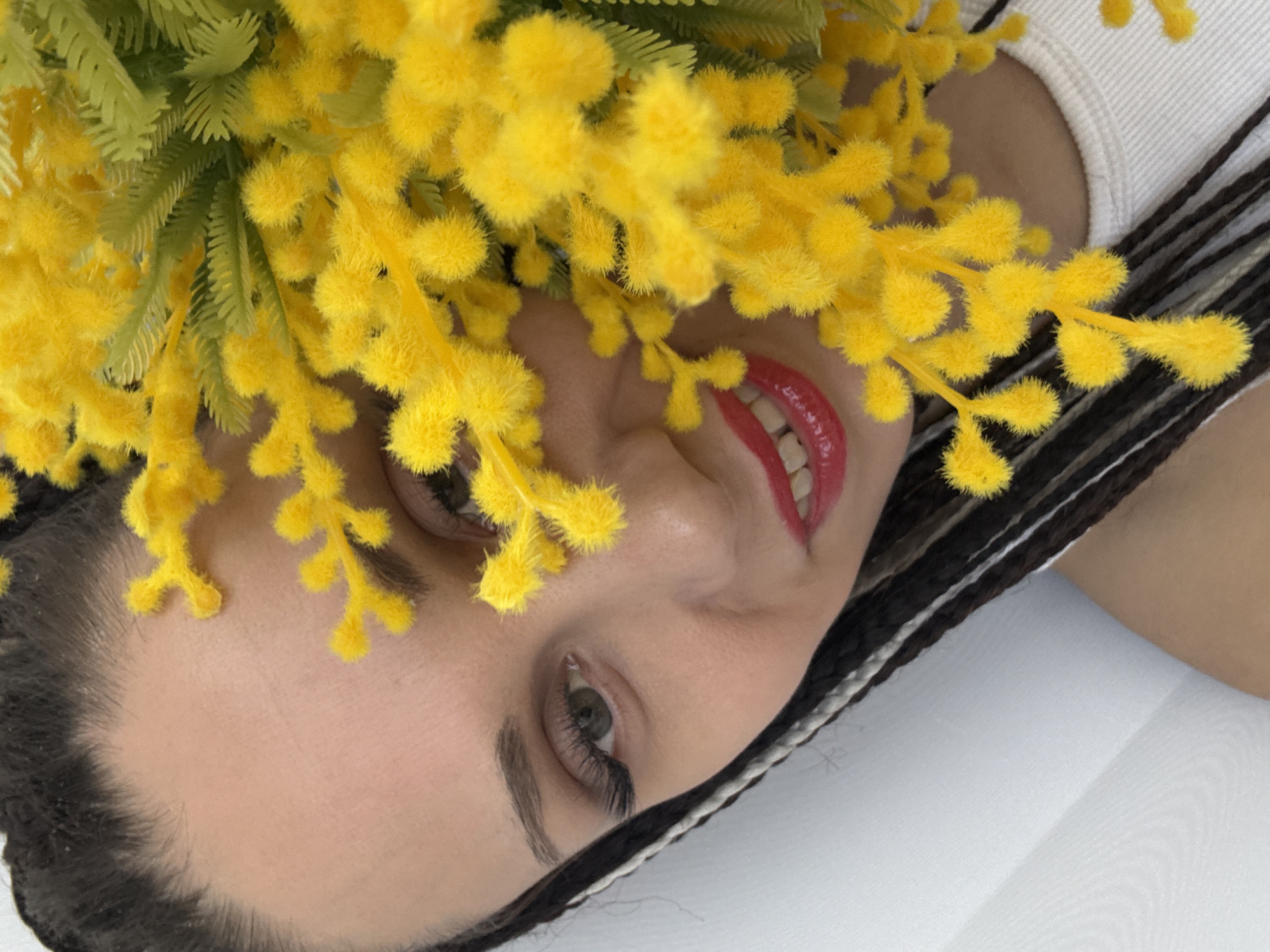 A close-up shot presents a person lying down, adorned with yellow mimosa flowers, with braided hair, against a white backdrop. The person has red lips and is wearing a white top.