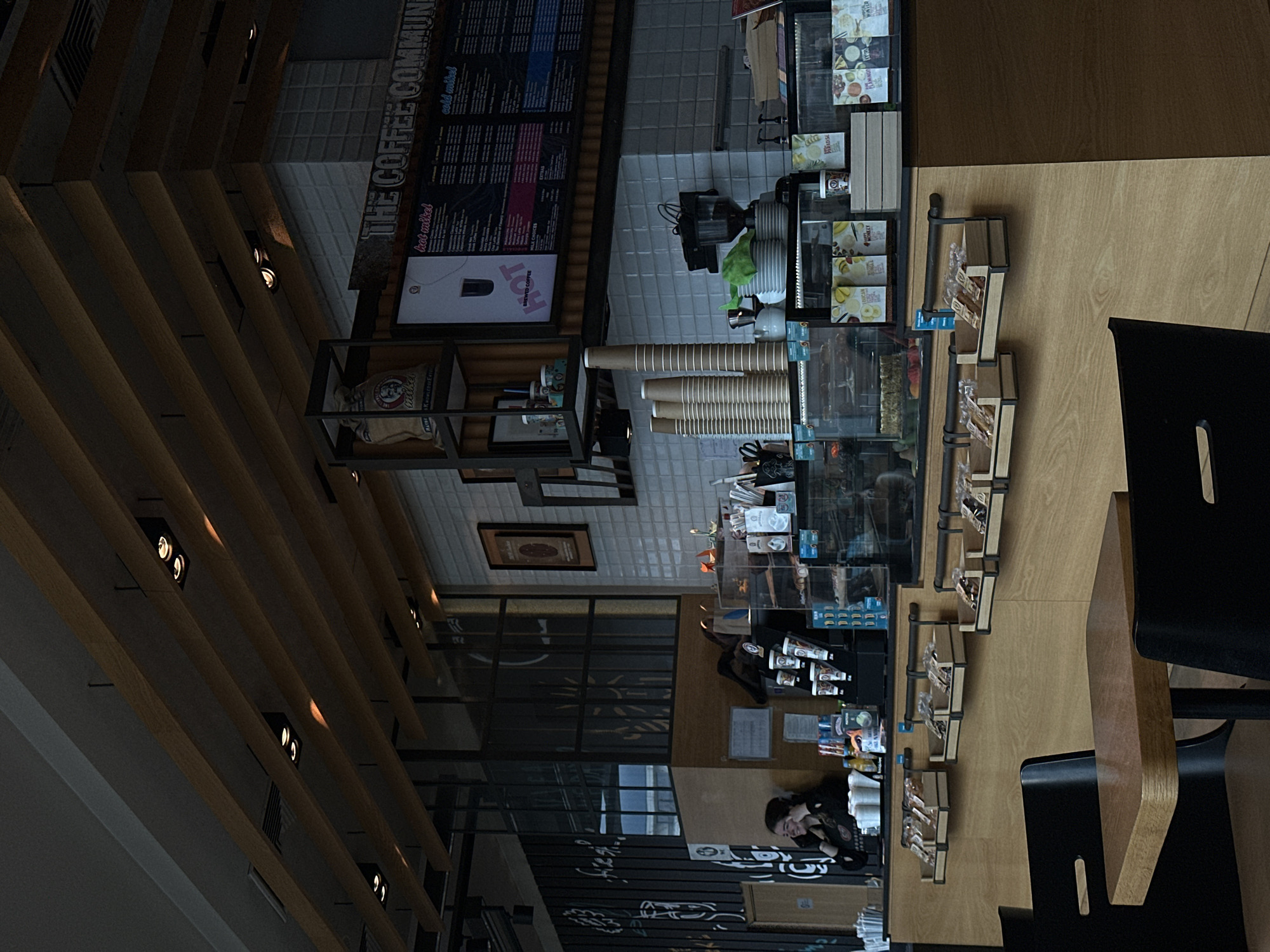 This is an interior shot of The Coffee Community, a cafe located in Sofia, Bulgaria. The perspective is from a seated position at a wooden table with black chairs. A person with long hair is visible at the service counter. The counter has glass-fronted display cases showcasing a variety of food items. Behind the counter are stacks of paper cups and coffee preparation equipment. A menu board with "THE COFFEE COMMUNITY" is mounted on the wall behind the counter. The ceiling is characterized by wooden slats with recessed lighting.