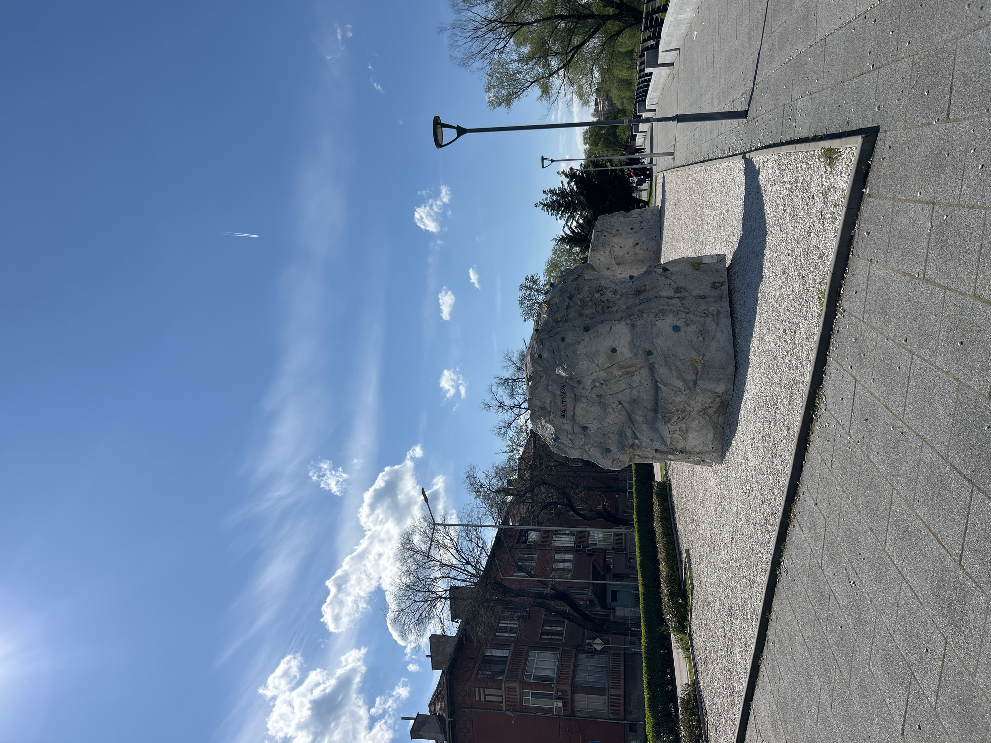 The image depicts an outdoor scene in Plovdiv, Bulgaria, featuring an artificial climbing boulder. The boulder is situated on a small area of gravel next to a paved walkway. A hedge separates the walkway from a multi-story residential building with red brick facades. In the background, there are trees and street lamps. The sky is blue with scattered clouds, and there is a contrail visible in the upper left portion of the frame.